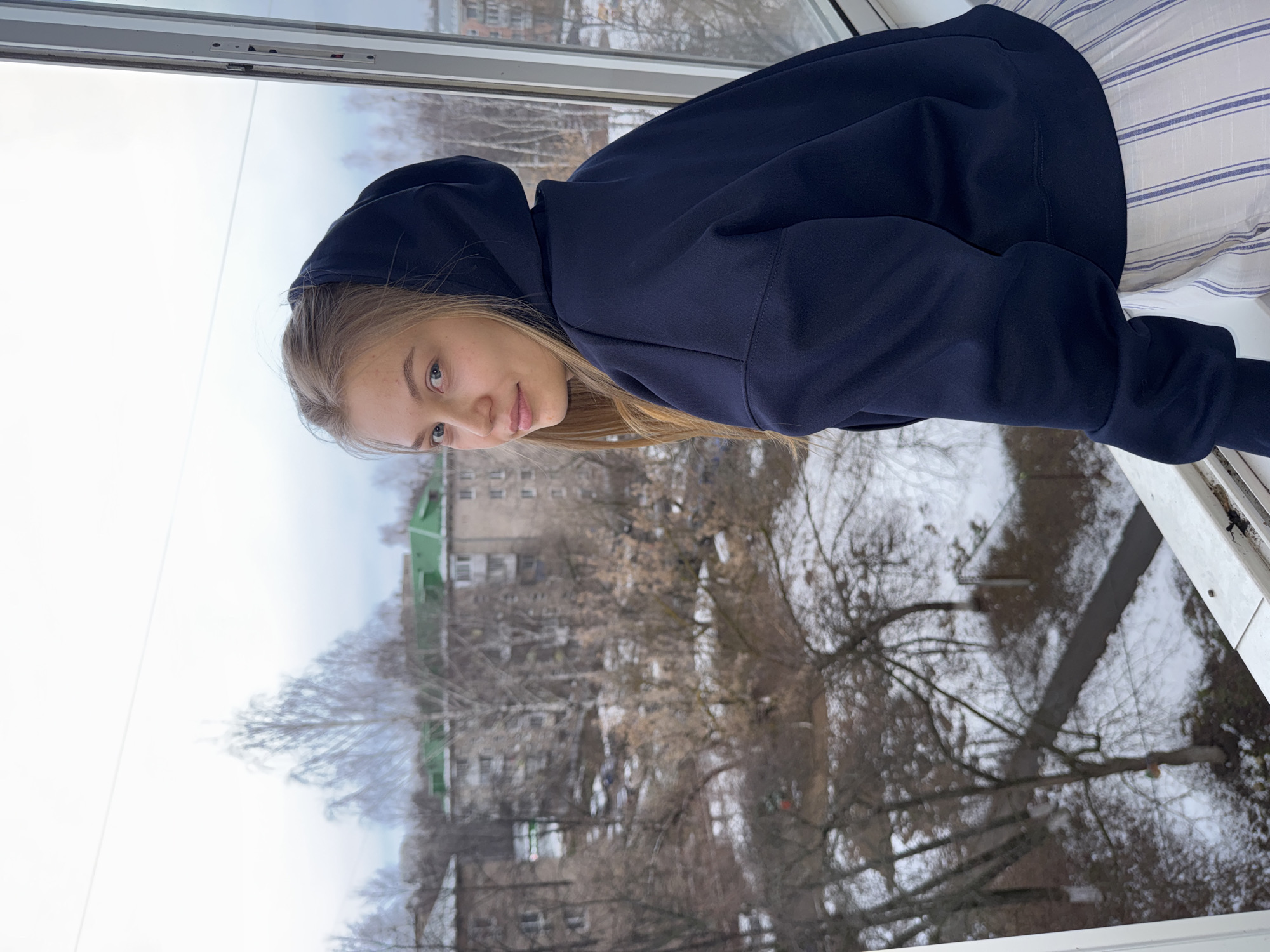 A young woman with blonde hair is sitting in front of a window. She is wearing a dark blue hoodie. Outside the window, bare trees and buildings are visible, suggesting an urban environment possibly in Chaikovsky, Russia. The sky appears overcast. The woman is looking at the camera. She appears to be indoors and is sitting on a striped surface.