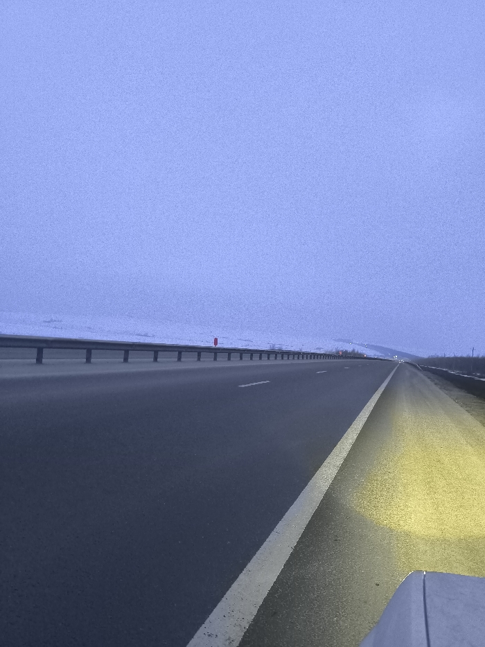 The scene depicts an empty multi-lane asphalt highway under a pale blue-grey, overcast sky, likely at dusk or dawn, in the vicinity of Pobeda, Russia. The viewpoint is from the front right of a vehicle, with a portion of its hood or bumper visible in the lower right quadrant, casting a bright yellow light beam onto the immediate right shoulder. The highway extends into the distance, flanked on the left by a metallic guardrail separating it from a snow-covered landscape of low hills or fields. A single red marker post is visible on the snowy embankment beyond the guardrail. The road surface is clear, marked with white dashed lane lines and a solid white line defining the right shoulder. No individuals or other vehicles are visible within the frame.