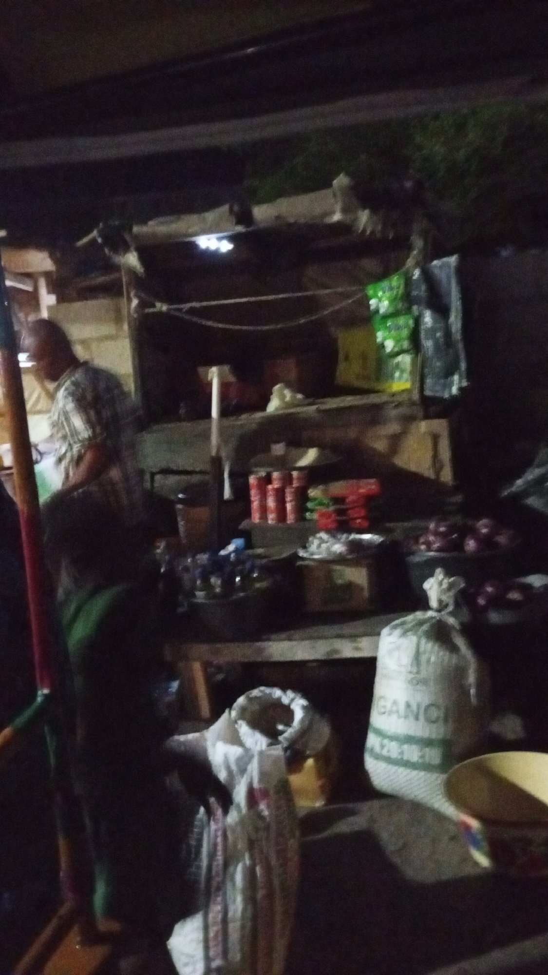 The image depicts an outdoor market stall in Adankolo, Nigeria, at night. The stall is constructed with wood and corrugated metal, illuminated by a single light bulb. A vendor wearing a patterned headscarf and a green garment stands behind the counter, possibly interacting with customers. Merchandise includes bags of grain or produce (one labeled "GANCI"), canned goods, packets of food, and onions. A bowl and other containers are visible on the ground in front of the stall. The surrounding environment is dark and indistinct.