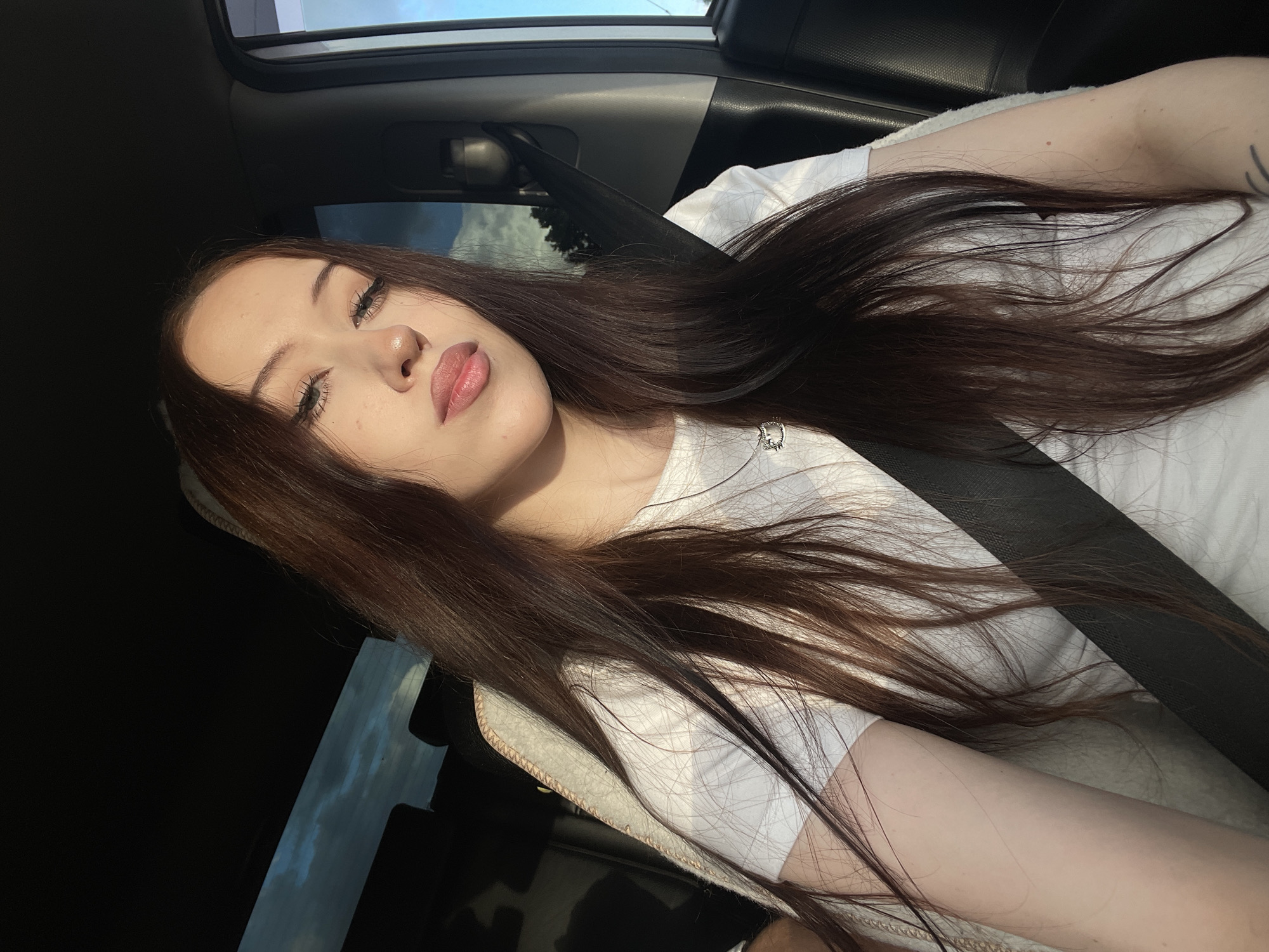 A young woman with long, dark brown hair is in the passenger seat of a car, taking a selfie. She is wearing a white t-shirt and a black seatbelt. The sun is shining, casting a warm glow on her face, and the sky is visible through the car window. Her eyes are closed, and her lips are slightly parted, suggesting a moment of relaxation or a playful pose. The scene appears to be set during the daytime, likely late afternoon due to the golden hour lighting. The background shows the interior of the car and a glimpse of the sky and trees outside, indicating she is likely on a road trip or a scenic drive. The overall mood of the image is relaxed and casual. No visible text is present in the image. The location context of Gavrilovka, Russia, does not provide specific visual cues within the image itself.