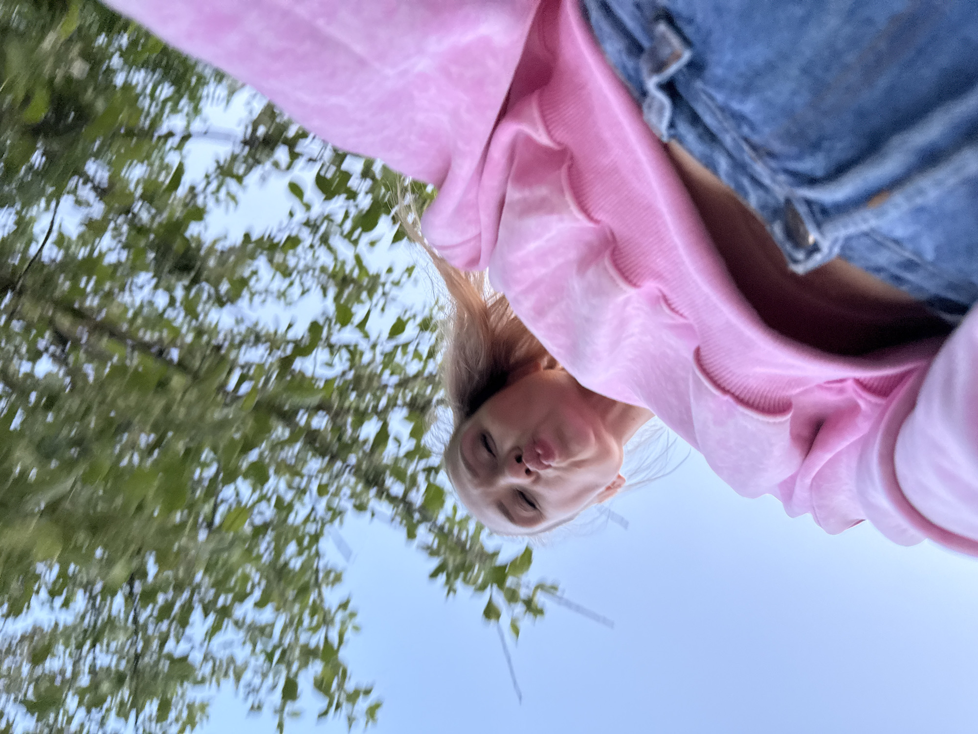 A photograph taken from a low angle depicts a person with blonde hair wearing a pink hooded sweatshirt and denim pants. The person is looking down at the camera with pursed lips. The background consists of green foliage on the left side and a clear, blue sky. The location, Chaikovsky, Russia, cannot be determined from the visual data provided.