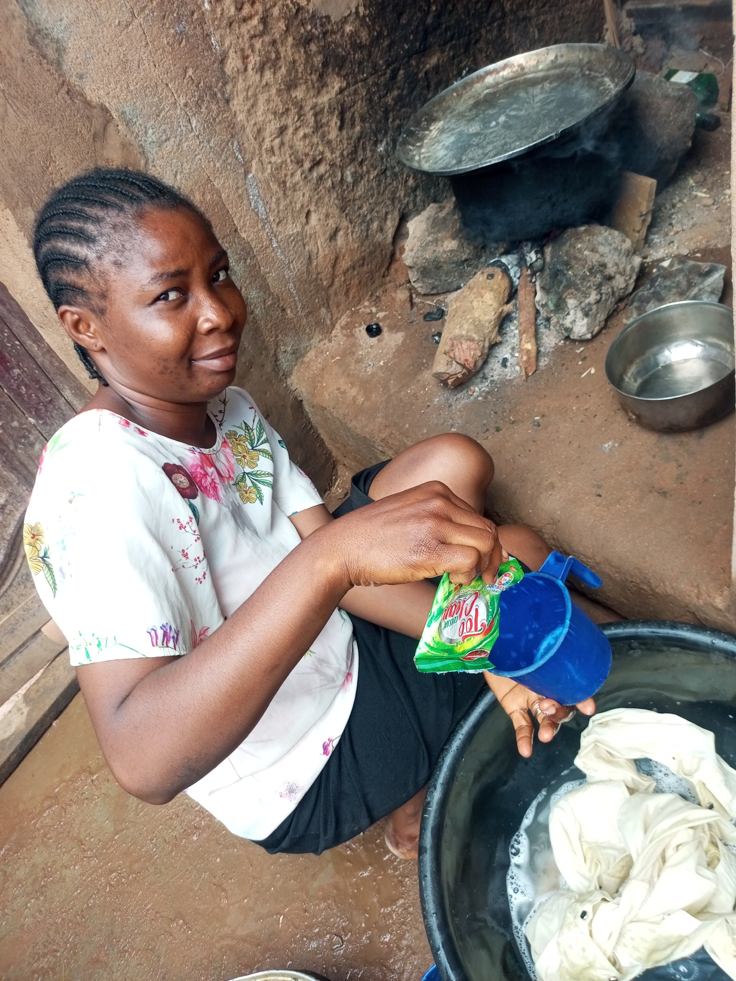 In Tempure, Nigeria, a Black woman with braided hair is captured in a medium shot, washing clothes in a black basin filled with soapy water. She is wearing a white blouse with a floral print. Her right hand holds a sachet labeled "Top Classic" and a blue plastic measuring cup, from which she appears to be pouring something into the basin. The background reveals a rustic setting with a clay wall, a cooking fire with a large metal pot over stones, and a smaller metal bowl to the right. The ground is made of packed earth, with some wet patches.