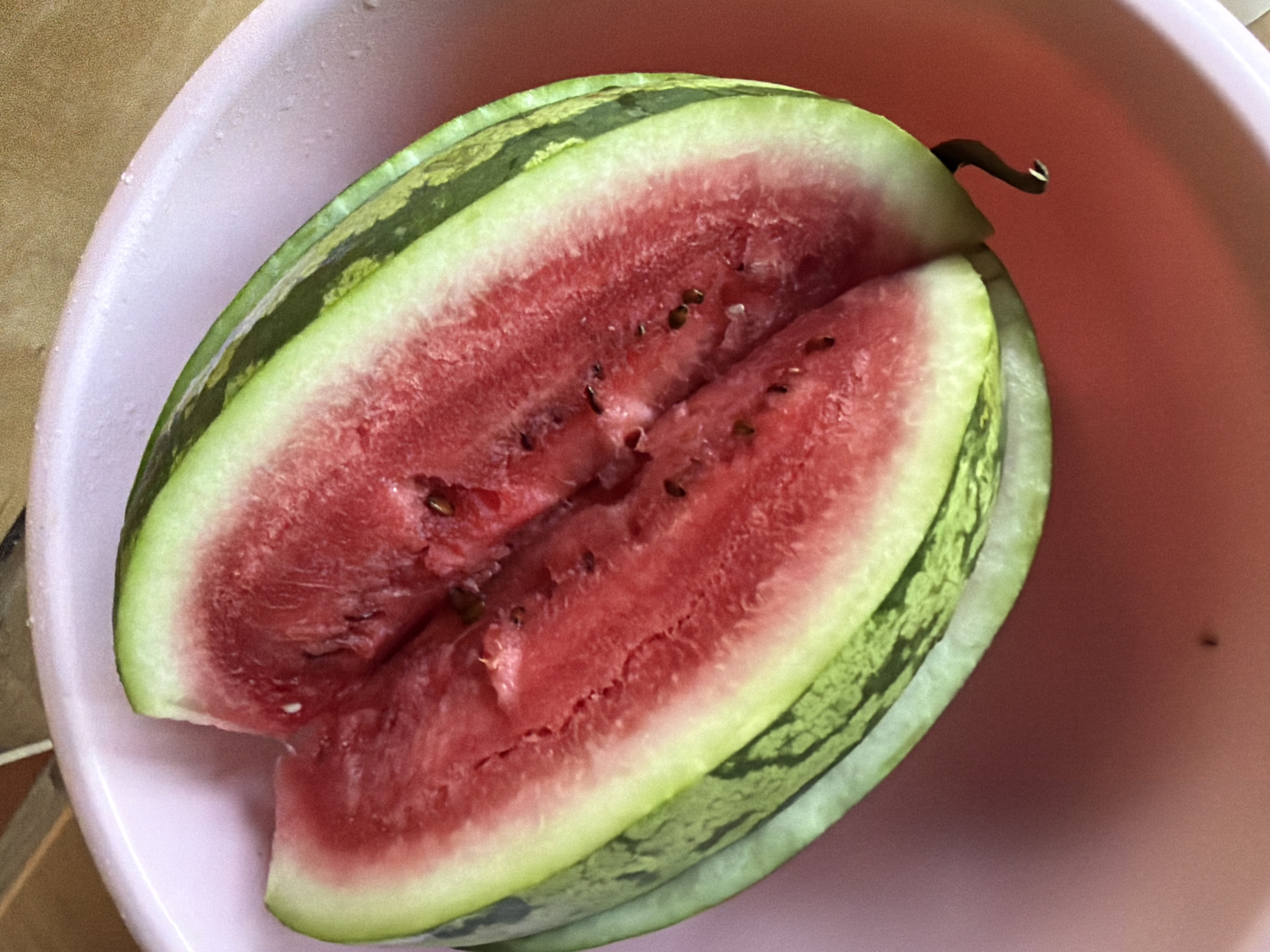 A close-up, overhead shot shows a cut watermelon resting in a pink plastic bowl. The watermelon is cut in half, revealing its bright red flesh filled with dark seeds. The rind is green with darker green stripes. The bowl is slightly tilted, with water droplets visible on its surface. The background is blurred, with textures suggesting a wooden surface and a wall. The scene is set in an indoor environment, with no specific city or location details available.
