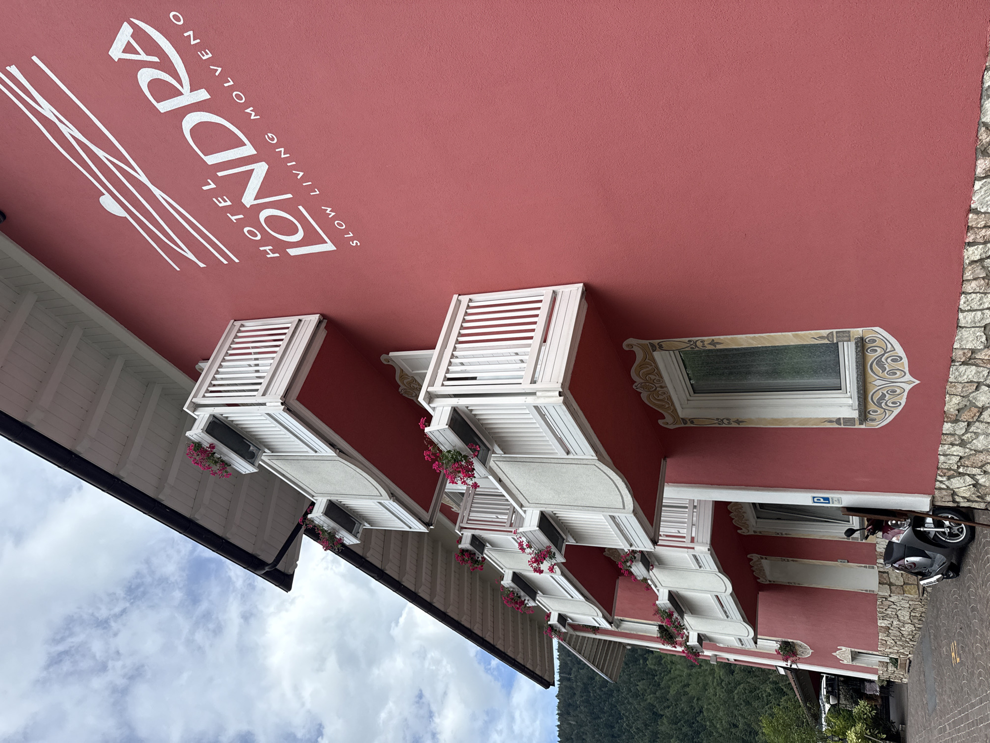 The image shows the exterior of the Hotel Londra in Molveno, Italy. The hotel has a bright pink facade with multiple white balconies adorned with pink flowers in planters. The hotel's name and slogan, "HOTEL LONDRA SLOW LIVING MOLVENO," are prominently displayed in white lettering on the wall. A scooter is parked in front of the hotel next to a stone-clad lower section of the building. In the background, a forested mountainside is visible under a cloudy sky.