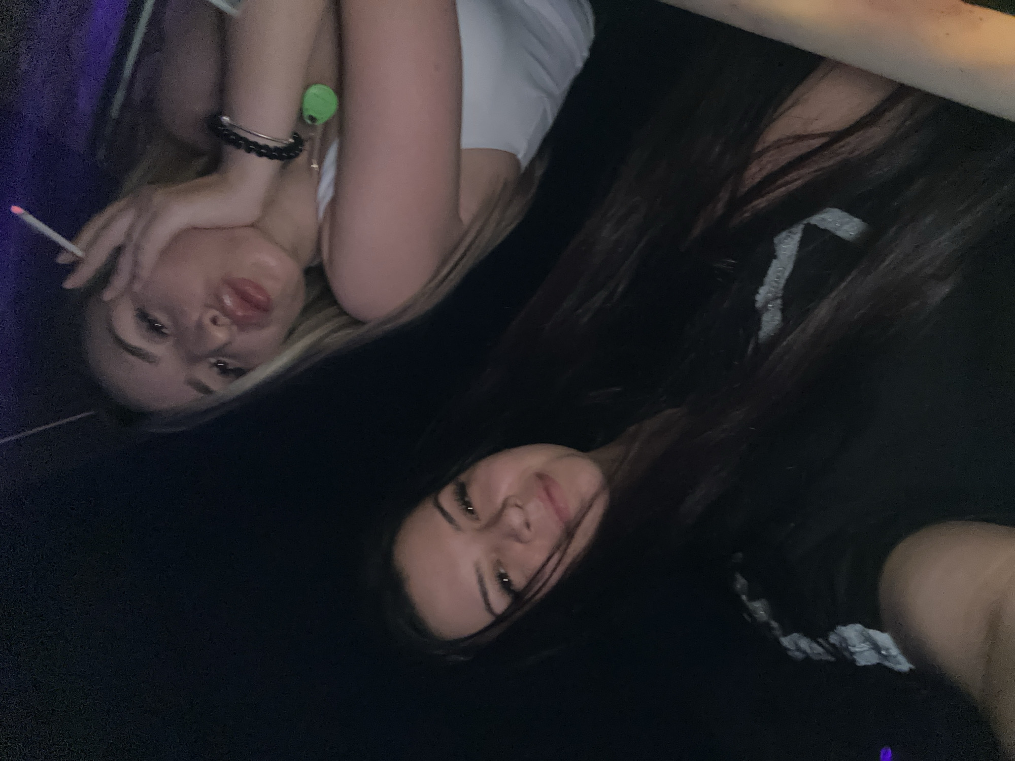 A low-light, slightly tilted vertical photograph features two young women in what is understood to be Sliven, Bulgaria.
The woman on the right, positioned in the upper portion of the frame, has blonde or heavily highlighted hair and is wearing a white tank top. Her left hand rests against her cheek, and she is holding a lit cigarette between her index and middle fingers. Her lips are slightly pursed, forming a "pout" expression. On her left wrist, she wears a black beaded bracelet, a metallic bangle, and a neon green, oval-shaped tag or accessory attached to a band. The background directly behind her upper right side shows a diffused purple light.
The second woman is positioned in the lower left portion of the frame, with her head and upper torso visible. She has long, dark hair, with some strands falling across her face, and is wearing a dark-colored top featuring a distinctive white, irregular, and slightly reflective pattern on the shoulder or upper arm area. She is smiling subtly, looking directly towards the camera.
The overall environment is dark, suggesting an indoor setting with artificial lighting, possibly at night.