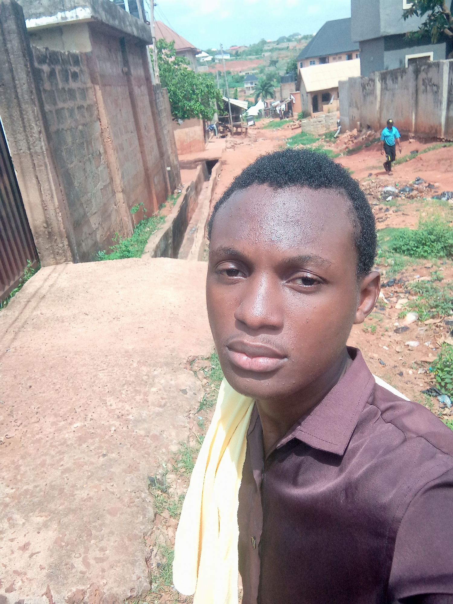 A young man takes a close-up selfie in an outdoor, semi-urban or rural setting in Ukwu-Abwa, Nigeria. He occupies the left foreground, looking directly at the camera with a neutral expression. He has short, dark, curly hair and appears to be perspiring on his forehead and face, suggesting warm weather. He wears a dark brown collared shirt, and a light yellow cloth is draped over his left shoulder.
Behind him, the scene depicts an unpaved, reddish-brown dirt road that slopes gently upwards into the distance. A concrete drainage ditch or canal runs alongside a tall, weathered concrete and brick wall on the left. Scattered green vegetation and weeds grow along the ditch and ground. In the mid-ground, a second man is seen walking away from the camera, wearing a light blue shirt and dark shorts.
Further in the background, several buildings of varying construction styles are visible, some appearing to be concrete block structures with tin or tiled roofs. The landscape includes distant hills with more buildings, suggesting a developing area. The bright, clear sky indicates daytime, likely midday or afternoon, consistent with the strong sunlight and short shadows. There is no visible text in the image.