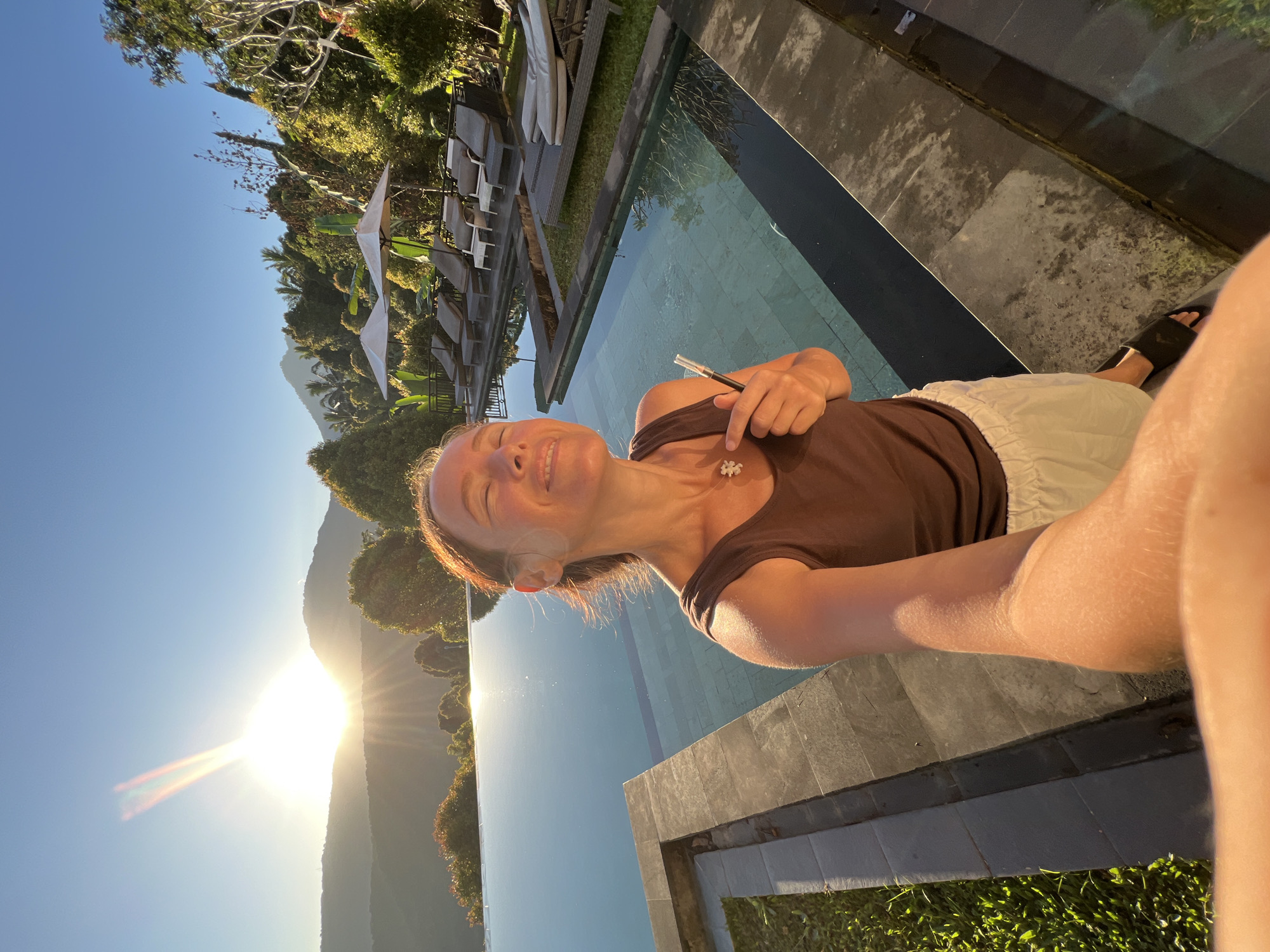 A woman is taking a selfie in front of an infinity pool at sunrise in Busungbiu, Indonesia. The sun is low on the horizon, casting a warm glow over the landscape. The woman is wearing a brown tank top and white shorts. She is smiling and has her eyes closed, enjoying the sun. The pool is surrounded by lush greenery and lounge chairs. The scene is serene and peaceful.