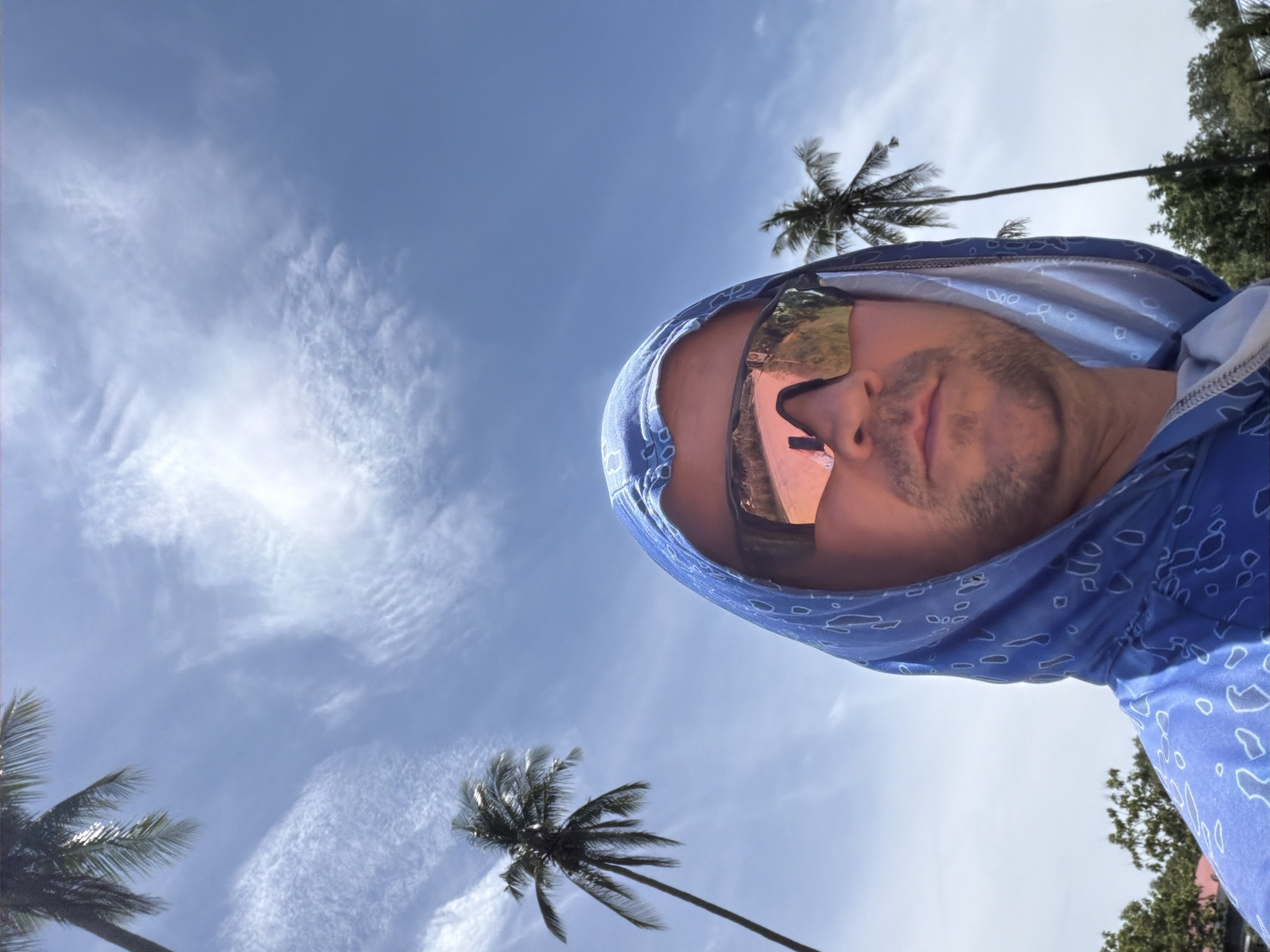 A man wearing sunglasses and a blue hooded jacket with a white pattern poses for a selfie under a bright blue sky with scattered clouds. Two palm trees are visible, one to the left and one to the right of the man. The man's face is partially obscured by his sunglasses, which reflect the surroundings. The overall impression is a sunny day in a tropical location, possibly Baan Bo Phut, Thailand, given the presence of palm trees and the clear sky.