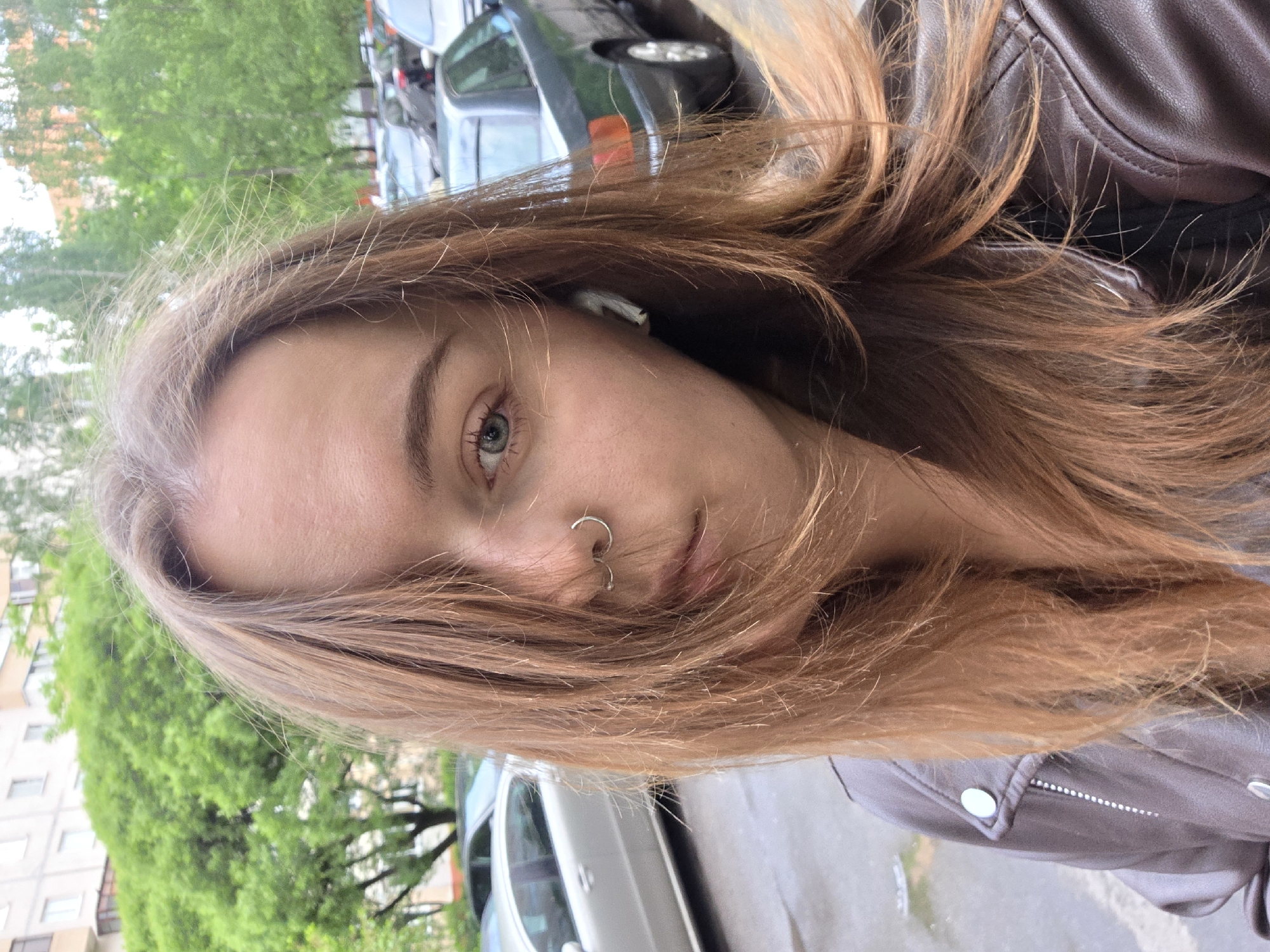 A close-up portrait shows a young woman with light brown hair, a nose ring, and light-colored eyes. She wears a brown leather jacket. The background appears to be an urban street scene with parked cars and green trees. The location appears to be a city street.