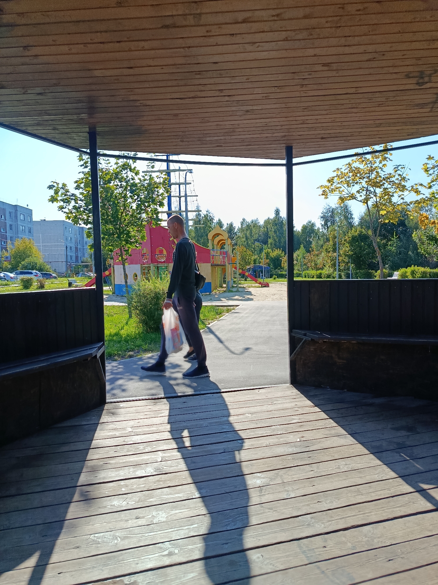 In the city of Vereya, Russia, a man walks out from under a wooden-planked shelter with a clear plastic bag in his right hand. He is wearing a dark long-sleeved shirt and dark pants. A second person's legs are visible behind him. The shelter has dark wooden benches on either side. Beyond the shelter, a children's playground with colorful structures is visible in the background, with trees and residential buildings further in the distance. The ground in front of the shelter is paved. Sunlight casts long shadows from the man and the shelter onto the wooden floor.