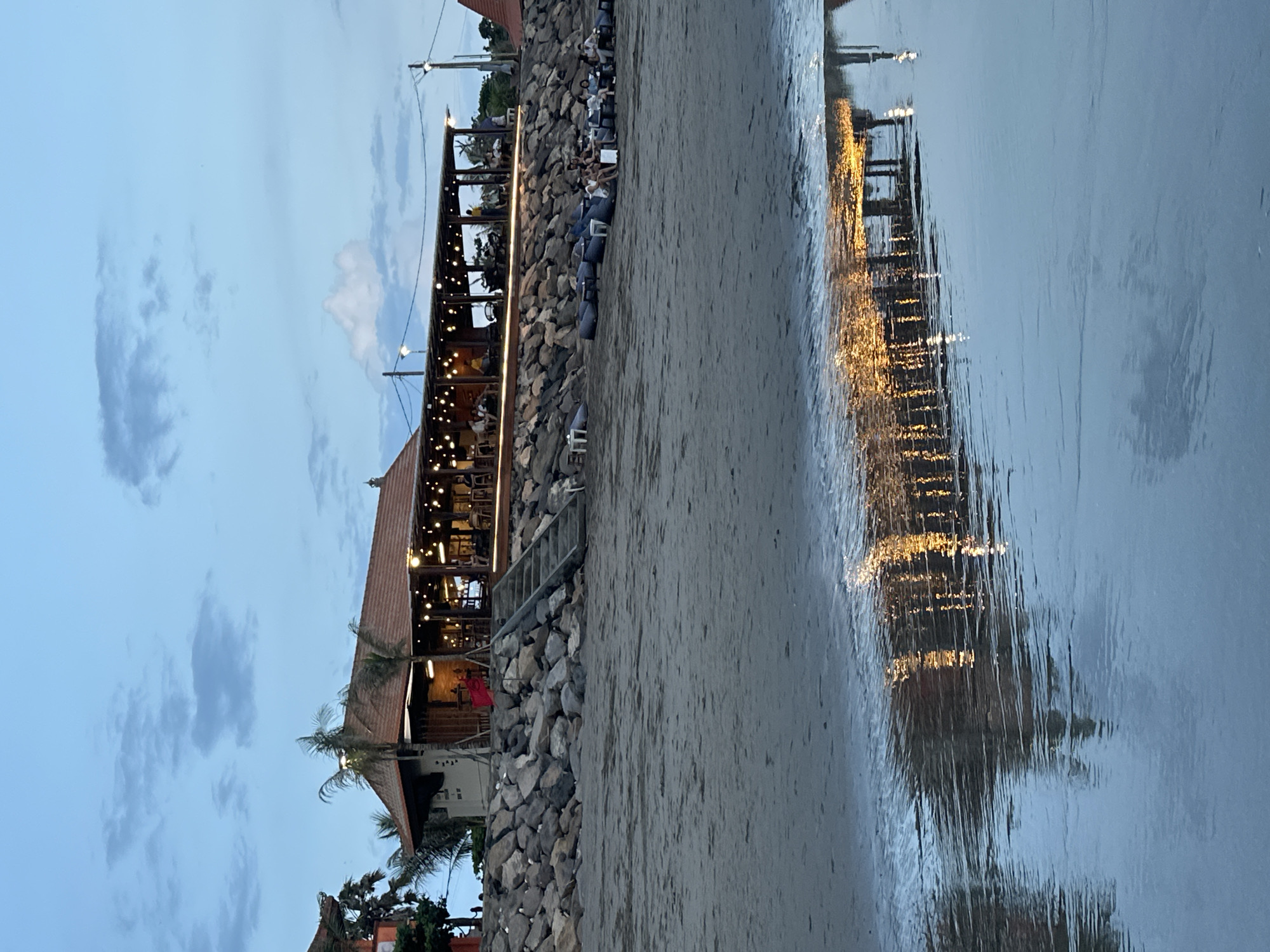 The image depicts a beachfront scene in North Kuta, Indonesia, likely at dusk or early evening. A multi-tiered building, potentially a restaurant or resort, stands parallel to the beach. The structure is constructed with wood and has a red tiled roof. The upper levels are open-air, illuminated by string lights and filled with people in sitting areas. The building sits on a rocky embankment which connects to the sandy beach. The foreground is comprised of sand and water, reflecting the lights from the building and creating a distorted, shimmering effect. The sky above is a gradient of blues and grays, suggesting the time of day. A few clouds are scattered across the sky. Vegetation, including palm trees, is visible near the building. People are visible on the upper floors of the building in the sitting areas.
