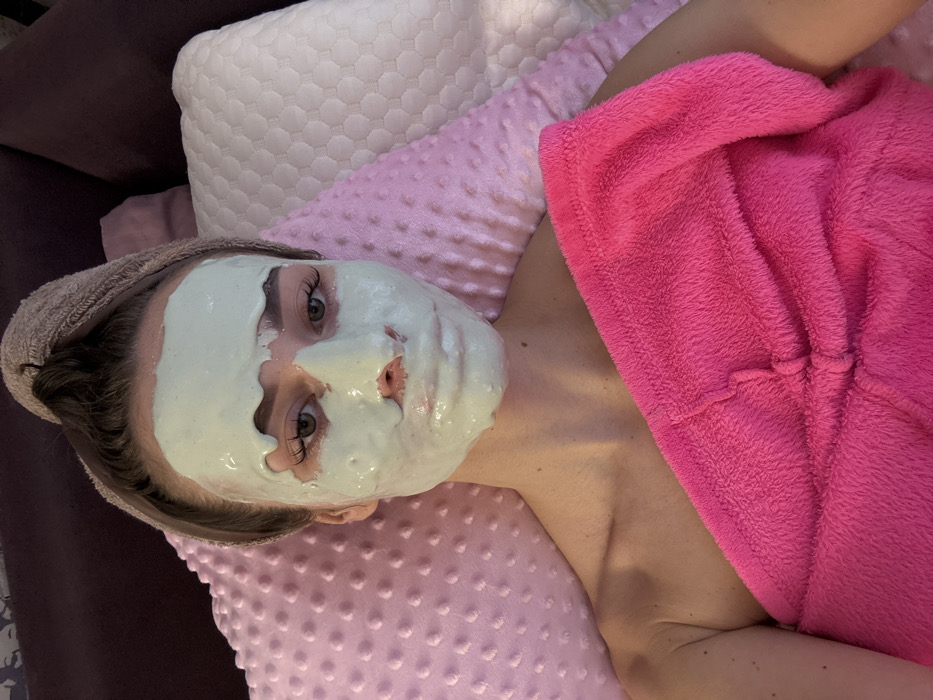 The image displays a female individual lying supine in an indoor setting. Her face is covered with a light green, viscous facial mask, leaving her eyes, eyebrows, and the area around her mouth exposed. Her hair is wrapped in a light brown terry cloth towel, twisted atop her head. She is partially covered by a bright pink, plush towel or wrap, which obscures her torso from the chest downwards, revealing her bare left shoulder and upper arm. She is situated against pillows; a light pink, textured pillow is to her left, and an off-white, hexagonally quilted pillow is to her right. A dark purplish-brown surface, likely upholstery or a headboard, forms the background. The specific location within Kyiv, Ukraine, or any other geographical area, cannot be determined from the visual information provided.