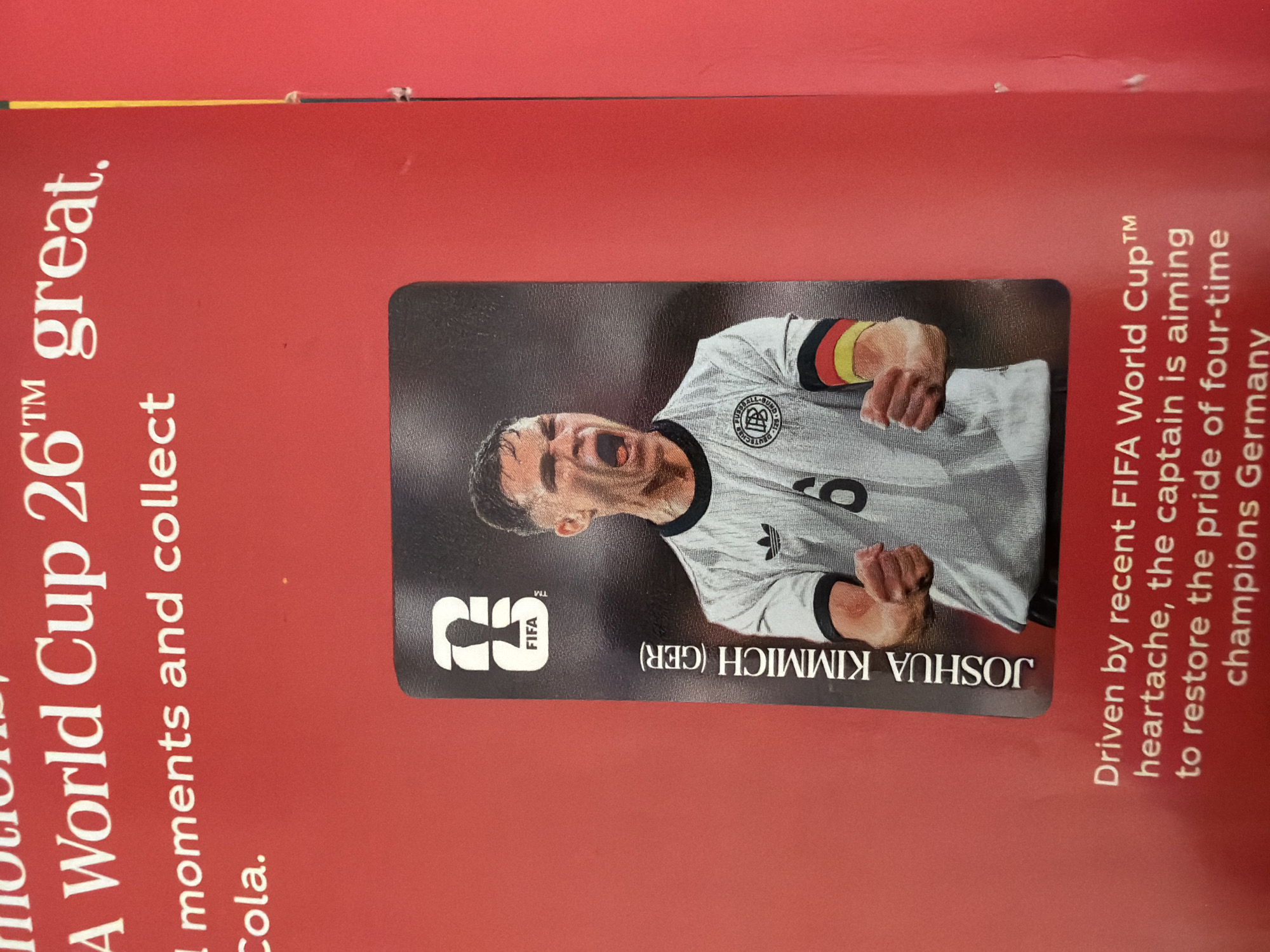 The image shows a red surface with text and a trading card. The text in white reads "A World Cup 26™ great. moments and collect Cola." Below this, a trading card features a soccer player, identified as "JOSHUA KIMMICH (GER)". The card also displays the FIFA logo. The player is wearing a white jersey with an Adidas logo and armbands in the colors of the German flag. The player is in a dynamic pose with his mouth open. The background is red, suggesting a promotional or display context. There is no visible indication of the city of Zlatitrap, Bulgaria, or any urban environment. The scene is a close-up of a printed surface and a trading card.