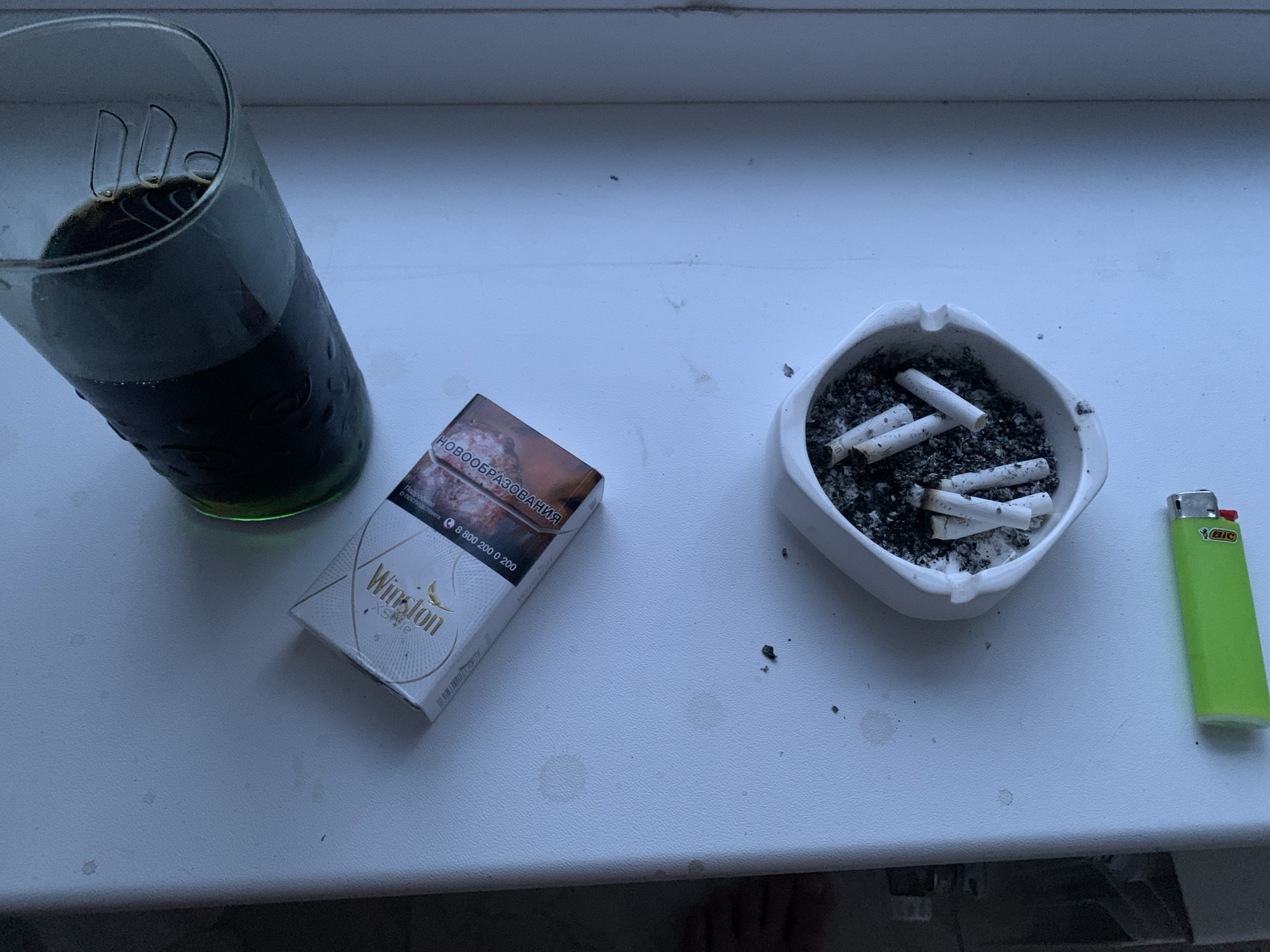 A top-down shot shows a still life on a white surface, likely a windowsill, in Stavropol, Russia. The scene is dimly lit, suggesting it is either early morning or late evening.
Dominating the left side is a tall glass filled with a dark liquid, possibly cola or coffee, with condensation on the outside and what appear to be ice cubes or remnants of them inside. To the right of the glass, a pack of Winston cigarettes is placed at an angle. The pack is white with gold accents and features an image of what looks like lungs, a common health warning. Text on the pack is visible, including "Winston" and what appears to be Russian warning text "НОВОГООБРАЗОВАНИЯ" (neoplasms) and a phone number "8 800 200 0 200".
Further to the right, a white ashtray contains several cigarette butts and ash, indicating recent use. Next to the ashtray lies a bright green BIC lighter. The white surface has a few faint circular stains, perhaps from condensation or spills. The overall mood is somber and possibly indicative of a moment of quiet reflection or a break.