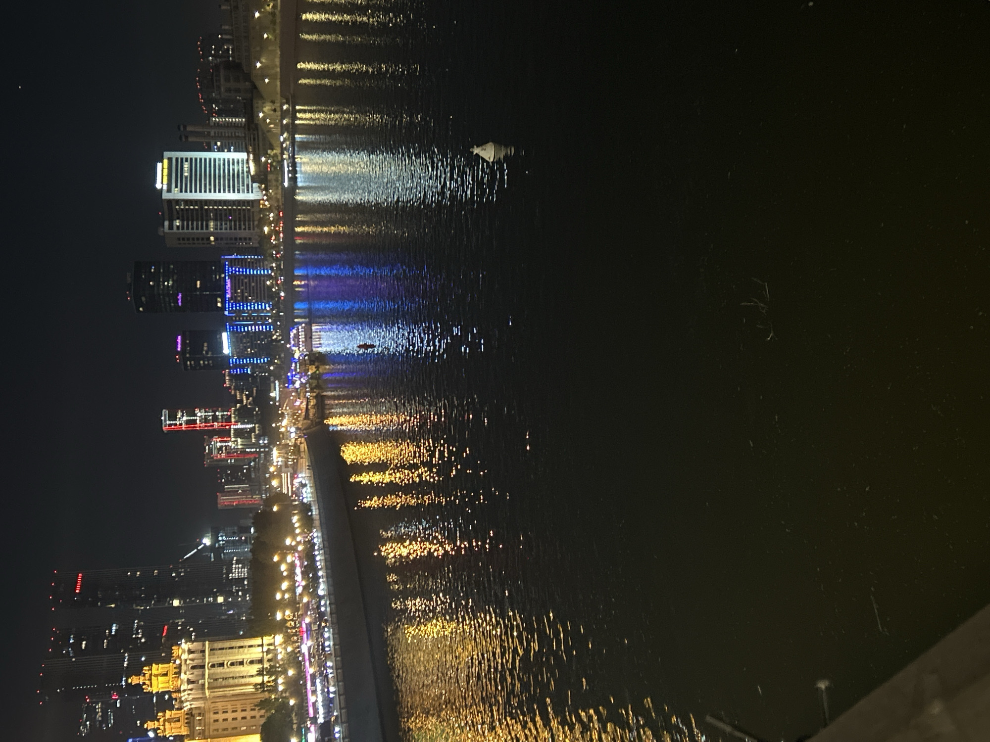 The image depicts a night scene of a city waterfront with multiple high-rise buildings illuminated with various colors. The lights reflect on the water surface, creating shimmering patterns. In the middle of the water, there is a small boat or floating object. The city's skyline is visible against the dark sky. Some elements near the foreground are blurred.