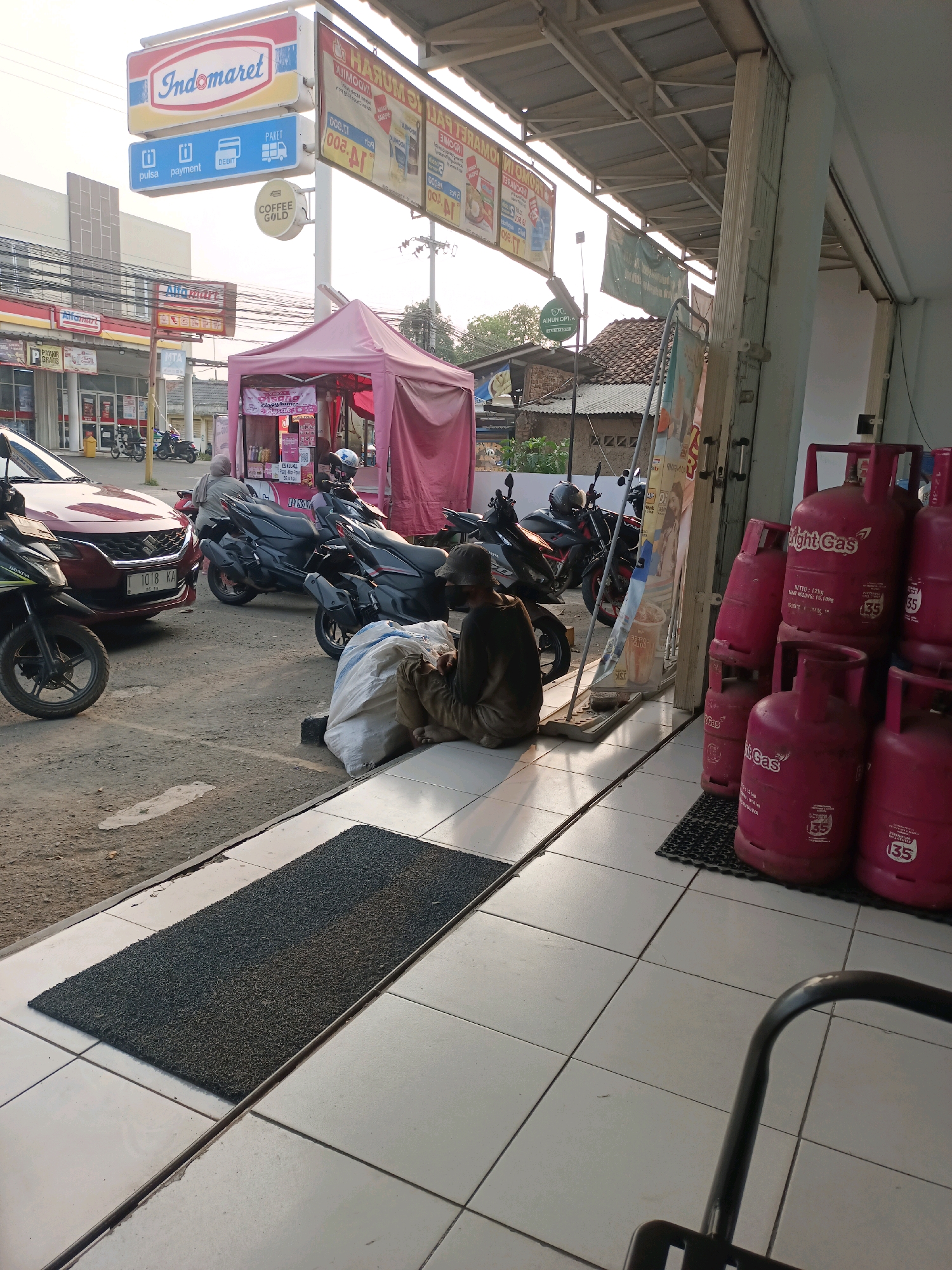 The image shows an outdoor scene in front of a convenience store, likely in Karawang, Indonesia, given the presence of "Indomaret" and "Alfamart" signage. In the foreground, a man wearing a dark long-sleeved shirt and brown pants is sitting on the ground, facing away from the camera. He is next to a large, white, lumpy sack. To his right, a stack of bright pink propane gas cylinders are lined up. The cylinders have yellow and white labels that read "Light Gas" and display "135" in a red circle. The ground is tiled with off-white squares. A black rubber mat is laid out on the tiles in front of the man and the gas cylinders.
In the midground, several motorcycles are parked. One is a dark red car. There are also pink and white stalls set up. In the background, more buildings and trees are visible under a bright, overcast sky. An "Indomaret" sign is prominently displayed on a pole with additional signs indicating payment options. Another sign for "Alfamart" is visible to the left. The overall impression is a busy commercial area with various shops and transportation.