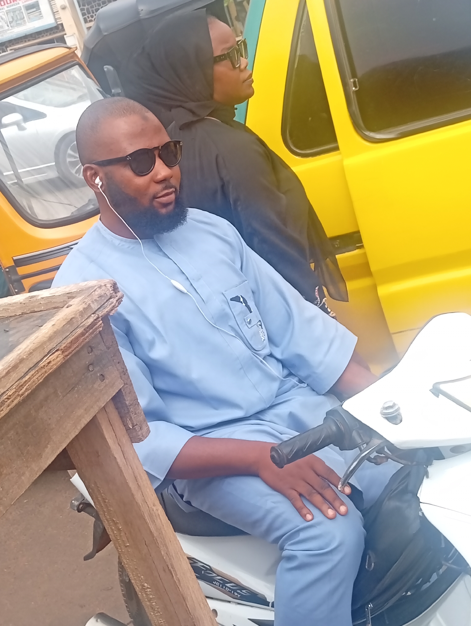 A man sits on a white motorcycle on a street in Maiburiji, Nigeria. He is wearing sunglasses, a blue robe, and white headphones. Behind him to the right is a woman wearing a dark headscarf and sunglasses, next to a yellow vehicle. A wooden structure is to the left of the man. The location appears to be a street in an urban setting.