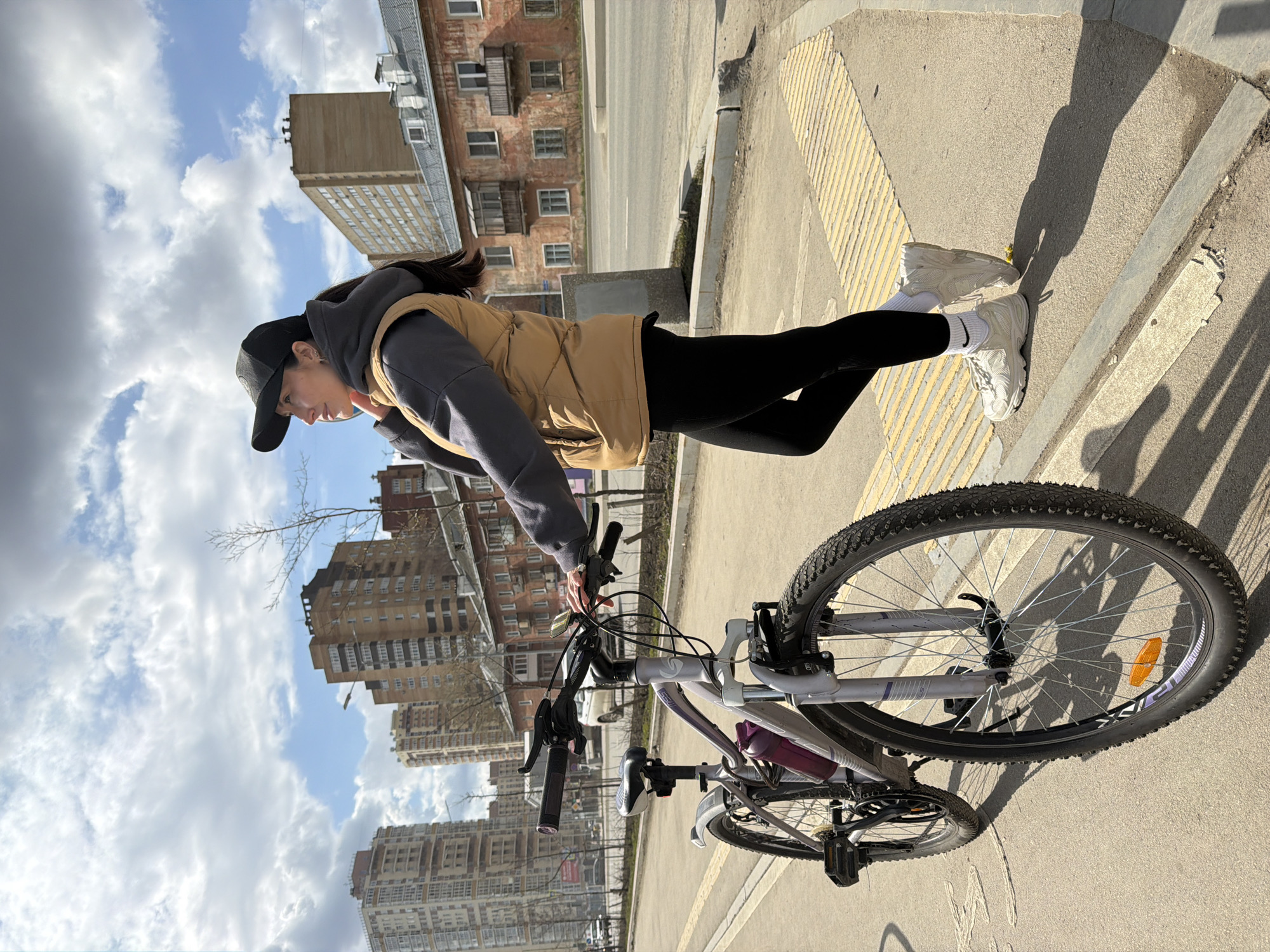 The image displays a young woman standing beside a bicycle on a paved urban street in Perm, Russia, under a partly cloudy sky.
The woman, appearing to be in her late teens or early twenties, has dark hair and is dressed in casual athletic wear: a black baseball cap, a dark grey hooded sweatshirt, a tan sleeveless puffer vest, black leggings, and white athletic shoes worn with white socks that feature a visible Nike logo. She is seen from the side, looking downwards, with one hand near the bicycle's handlebars.
The bicycle is a light grey mountain or hybrid bike with knobby tires, a front reflector, and a dark purple water bottle secured in a cage on its frame. The setting includes a paved sidewalk or street with a concrete curb and distinctive yellow textured tactile paving, likely indicating a pedestrian crossing or ramp.
In the background, there are various multi-story urban buildings. Some appear to be older, constructed of brick with balconies, while others are taller, more modern apartment blocks. Bare tree branches are visible against the sky in some areas. Shadows cast on the ground by the woman and the bicycle suggest it is daytime, possibly mid-day or afternoon. The sky is partly covered with white and grey clouds. The overall scene depicts a casual moment of a person with their bicycle in a city environment.