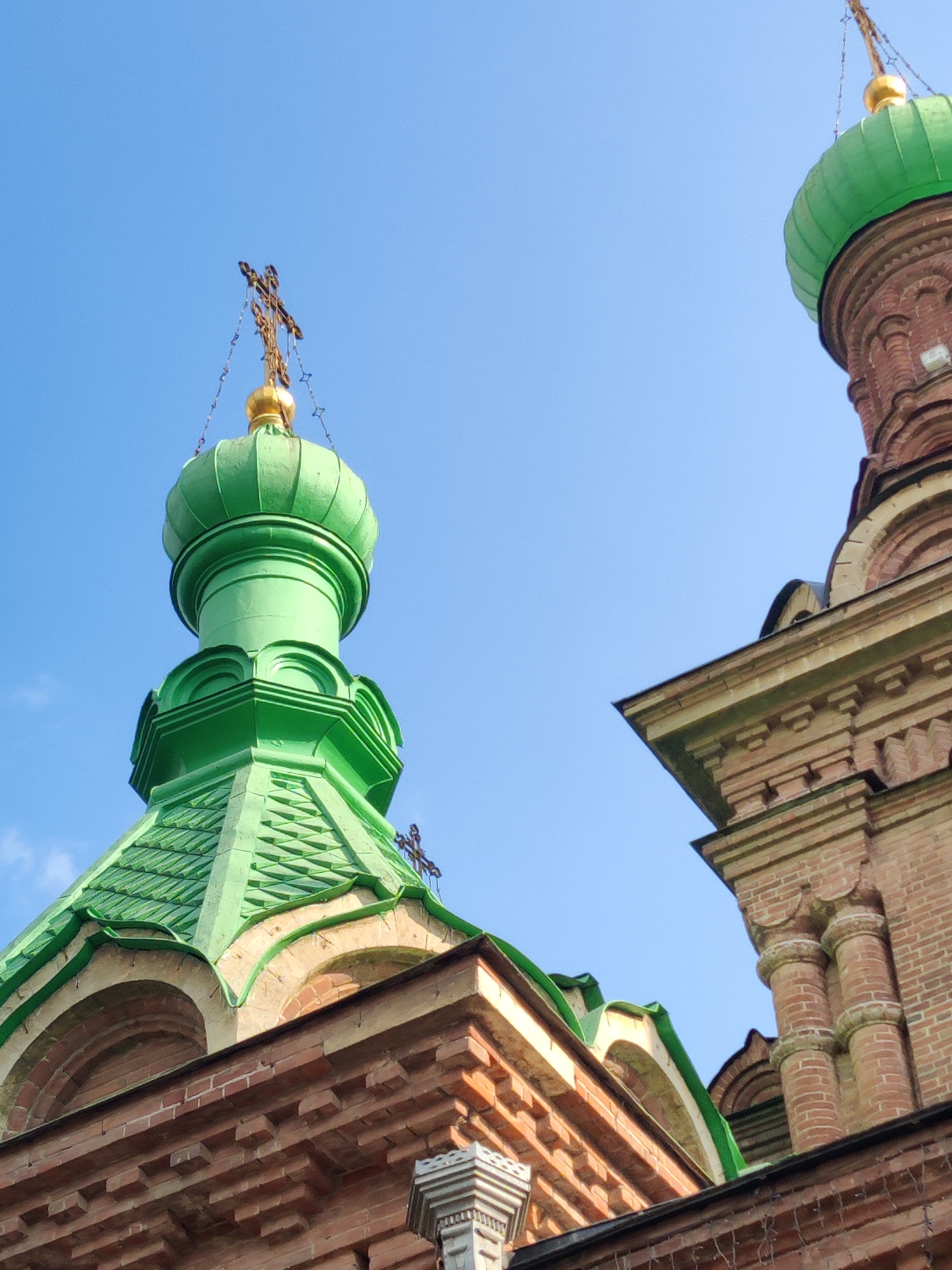 The image is a low-angle shot of two church domes against a clear blue sky. The dome on the left is predominantly green, with a golden finial topped by a cross. It is made of metal with ribbed detailing. The dome on the right is also green and has a similar golden finial and cross, but it is partially obscured by the brick structure of the church. The churches are constructed of red brick with architectural details such as arched windows and decorative brickwork. The perspective suggests the photo was taken from the ground looking up. This is a view of the Cathedral of St. Catherine in Krasnodar, Russia.