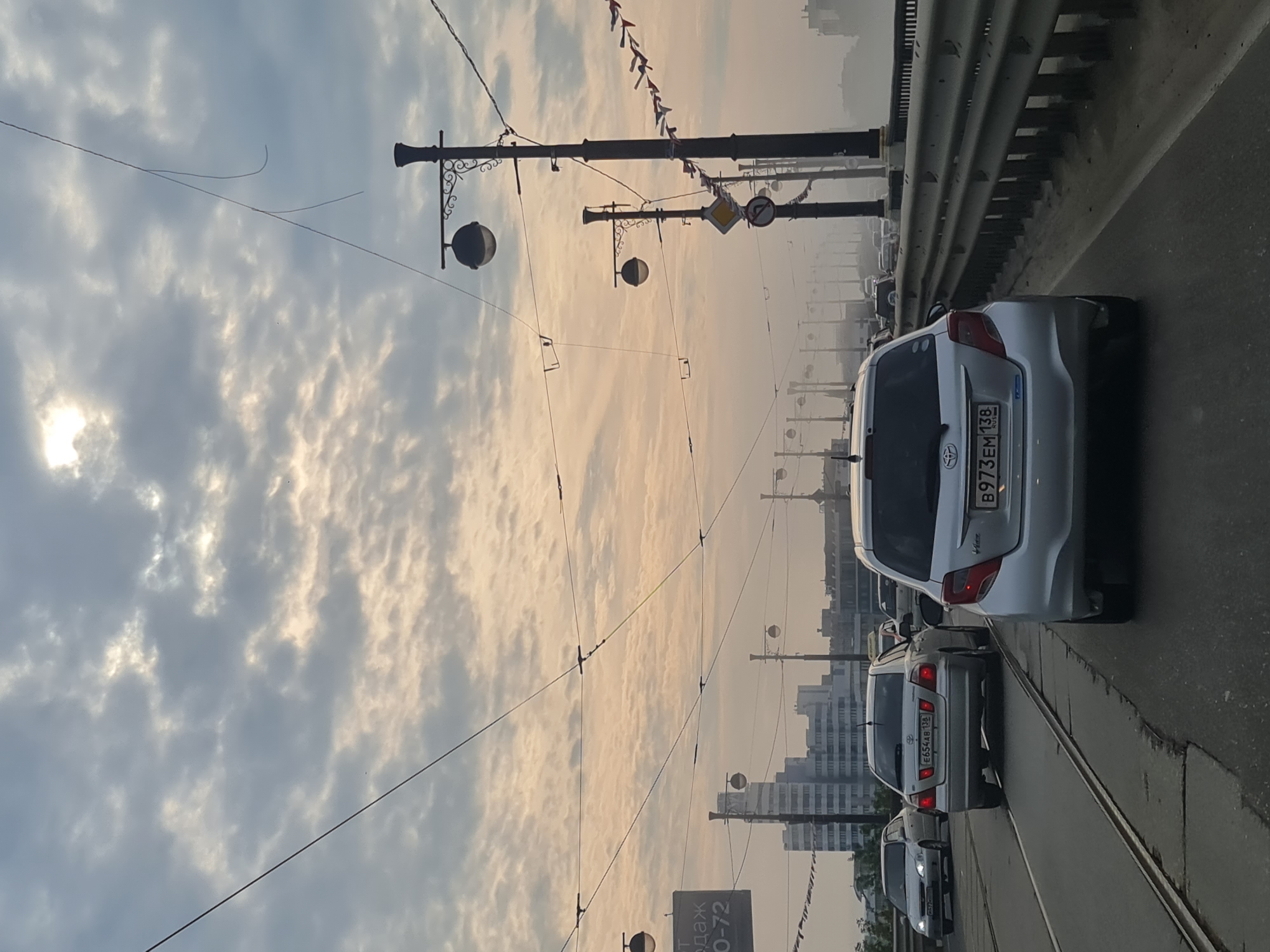 The image depicts a cloudy sky over a bridge with several cars driving on it. The sky is filled with a mix of gray and white clouds, with the sun faintly visible through a patch of brighter clouds. Overhead wires crisscross the sky, supported by lampposts. In the foreground, a silver hatchback car is seen from the rear. To its left, another silver sedan is driving, and further ahead, a white sedan is visible. A city skyline with tall buildings can be seen in the distance, partially obscured by a light haze. The bridge has a metal guardrail on the right side. The overall scene suggests a traffic situation on a bridge, possibly during the early morning or late afternoon given the lighting.
This scene is located on the **New Angara Bridge (Novyy Angarskiy Most)** in Irkutsk, Russia, which spans the Angara River. The distinctive architecture of the buildings in the background, specifically the tall, Soviet-era style building on the left side of the image, is characteristic of Irkutsk. The presence of tram tracks embedded in the road surface further confirms the location, as the New Angara Bridge is a major thoroughfare that carries tram lines in addition to vehicular traffic. The vehicles visible, including the Toyota hatchback and sedan, are common models seen in Russia. The Russian license plates on the cars (e.g., E654AB 138, B973EM 138) are also consistent with the location. The signage on the lampposts, though partially obscured, likely contains Russian text.