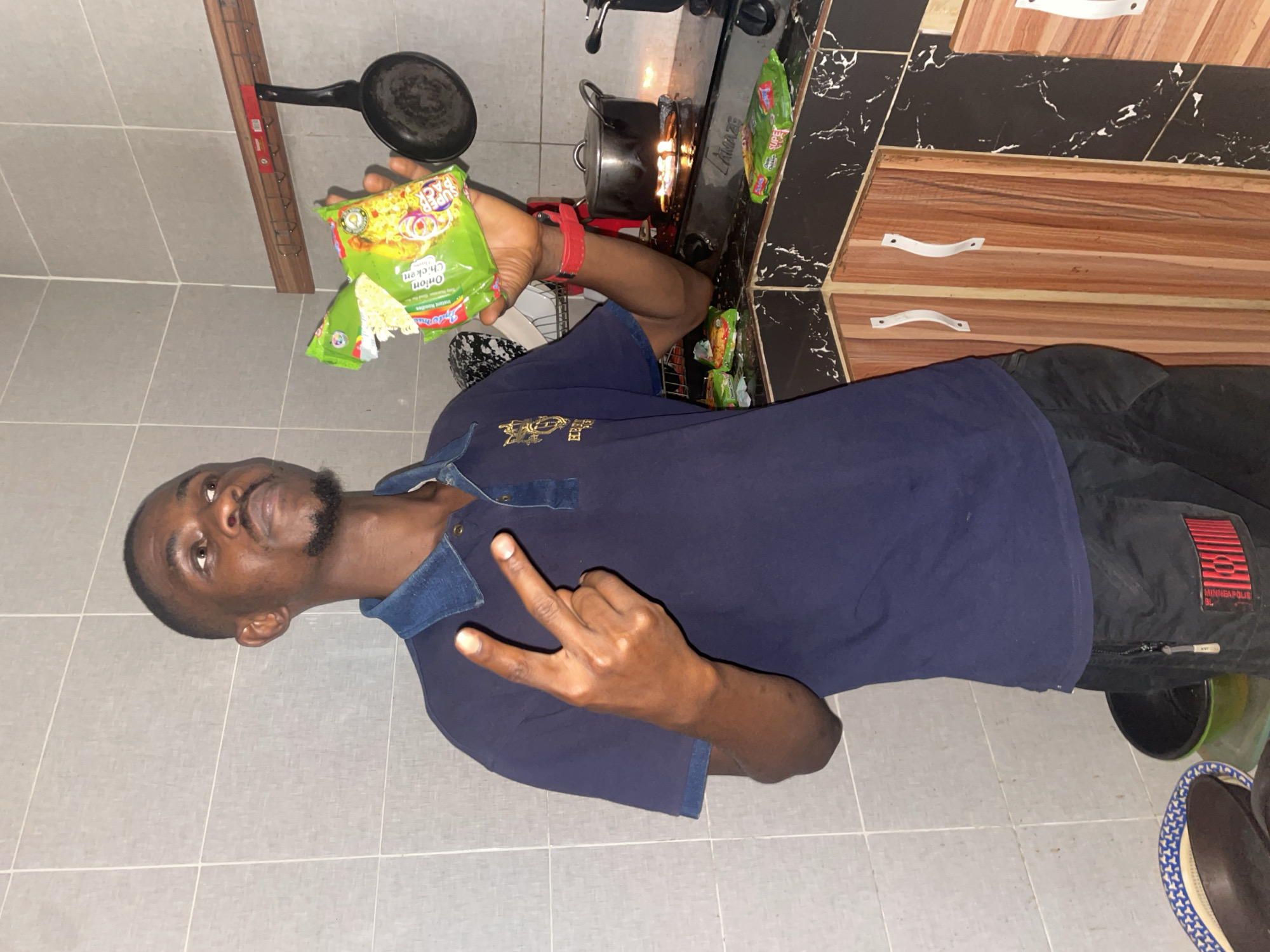 An adult male is holding a packet of Indomie Onion Chicken flavor instant noodles with the packaging torn open in his left hand. His right hand is raised, making a "W" or "west side" hand gesture. He is wearing a dark blue polo shirt and black pants. A red wristband is visible on his left wrist. He is standing in a kitchen with gray floor tiles. Behind him is a black and white marble-patterned countertop. A gas stove with a pot on it is visible, with flames burning beneath the pot. Several unopened Indomie packets are on the countertop. A frying pan is hanging above the stove. There are light brown wooden cabinets with white handles above the countertop. It is not possible to ascertain the location specifically within the city of Abuja, Nigeria, based on visual information in the image.
