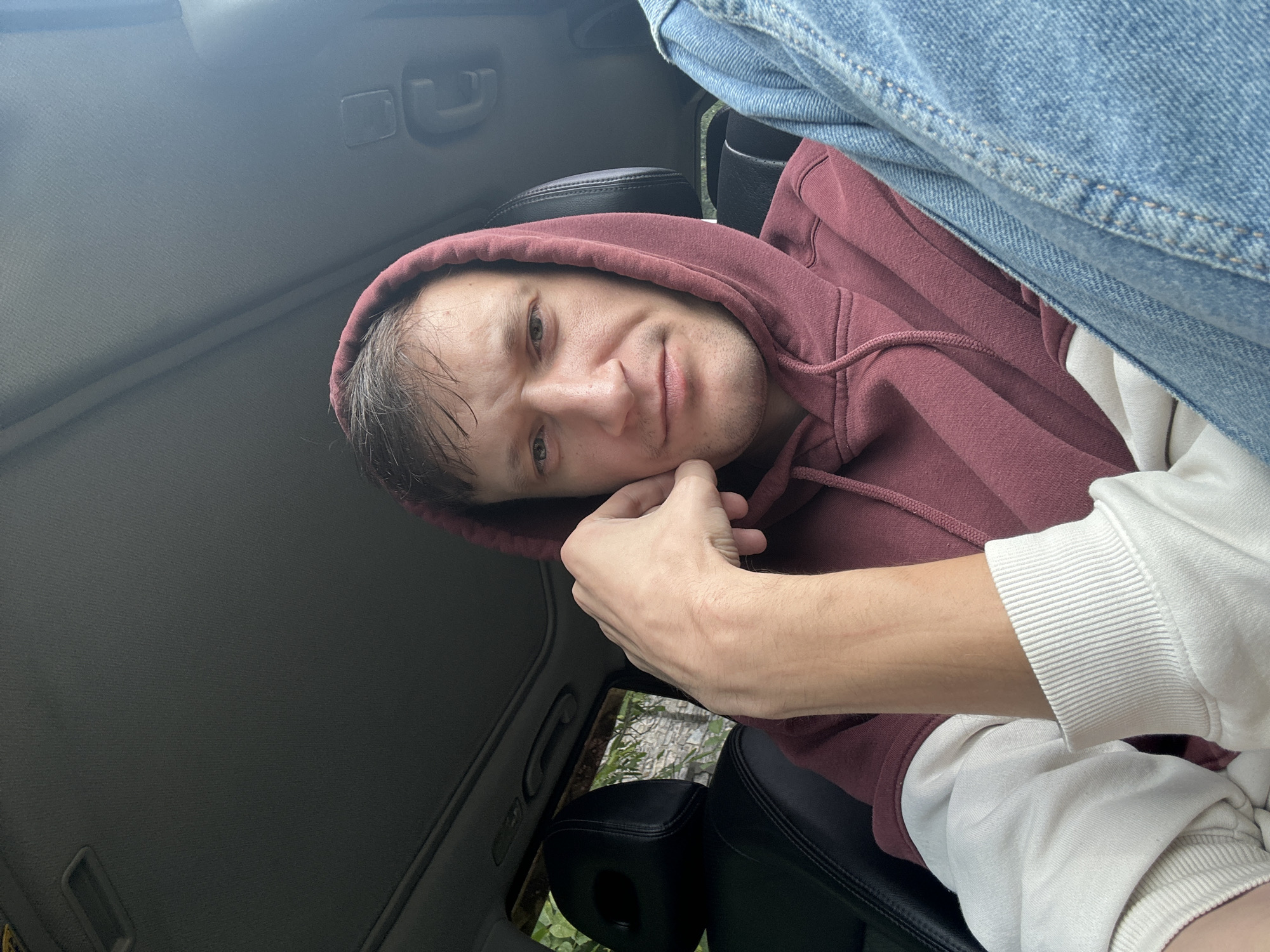 A man with light brown hair is sitting in the driver's seat of a car, resting his chin on his hand. He is wearing a maroon hooded sweatshirt with cream-colored sleeves and denim jeans. The car's interior is visible, including the headrest of the passenger seat and the sunroof. Through the window, trees and stone structures are visible, suggesting an outdoor, possibly rural or suburban setting, consistent with Makiv, Ukraine.