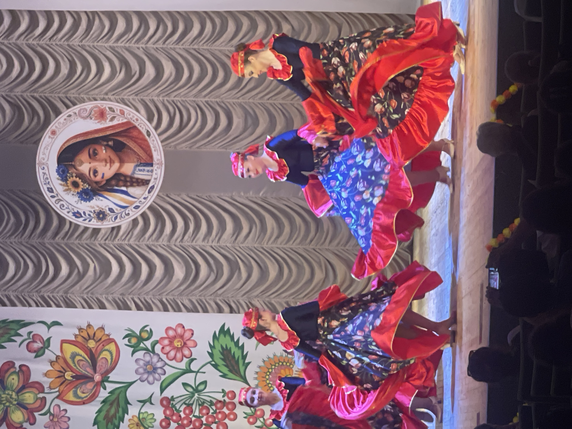 Four female dancers in traditional Ukrainian attire are performing on a stage in Гожули, Ukraine. The dancers are wearing dark blue tops with ruffled red collars and sleeves, and long, full skirts patterned with flowers and trimmed with red ruffles. They also wear matching red headscarves. The stage backdrop consists of draped curtains. To the left of the stage is a decorative panel with floral motifs, and a circular image of a young woman's face is positioned above the curtains. In the foreground, the audience is visible, with some individuals taking photos with their phones.