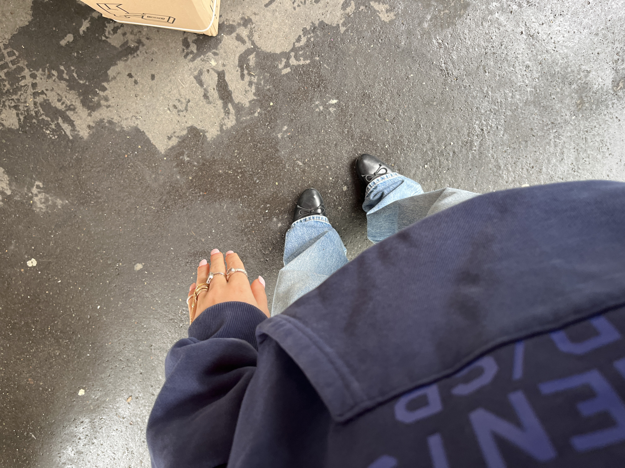 A ground-level, point-of-view image shows a person standing on a wet, mottled gray surface that appears to be concrete or asphalt. The person is wearing blue jeans and black ballet flat shoes with small bows. On the left hand, multiple rings are visible on the fingers. The person is wearing a dark blue hooded sweatshirt, partially obscuring the view, with large lettering on the lower portion. Part of a cardboard box is visible in the upper left corner. Based on the typical architectural style and the wet ground surface, it is possible that this image was taken in an urban area. Without more context, it is not possible to confirm if the scene is indeed Paris, France.