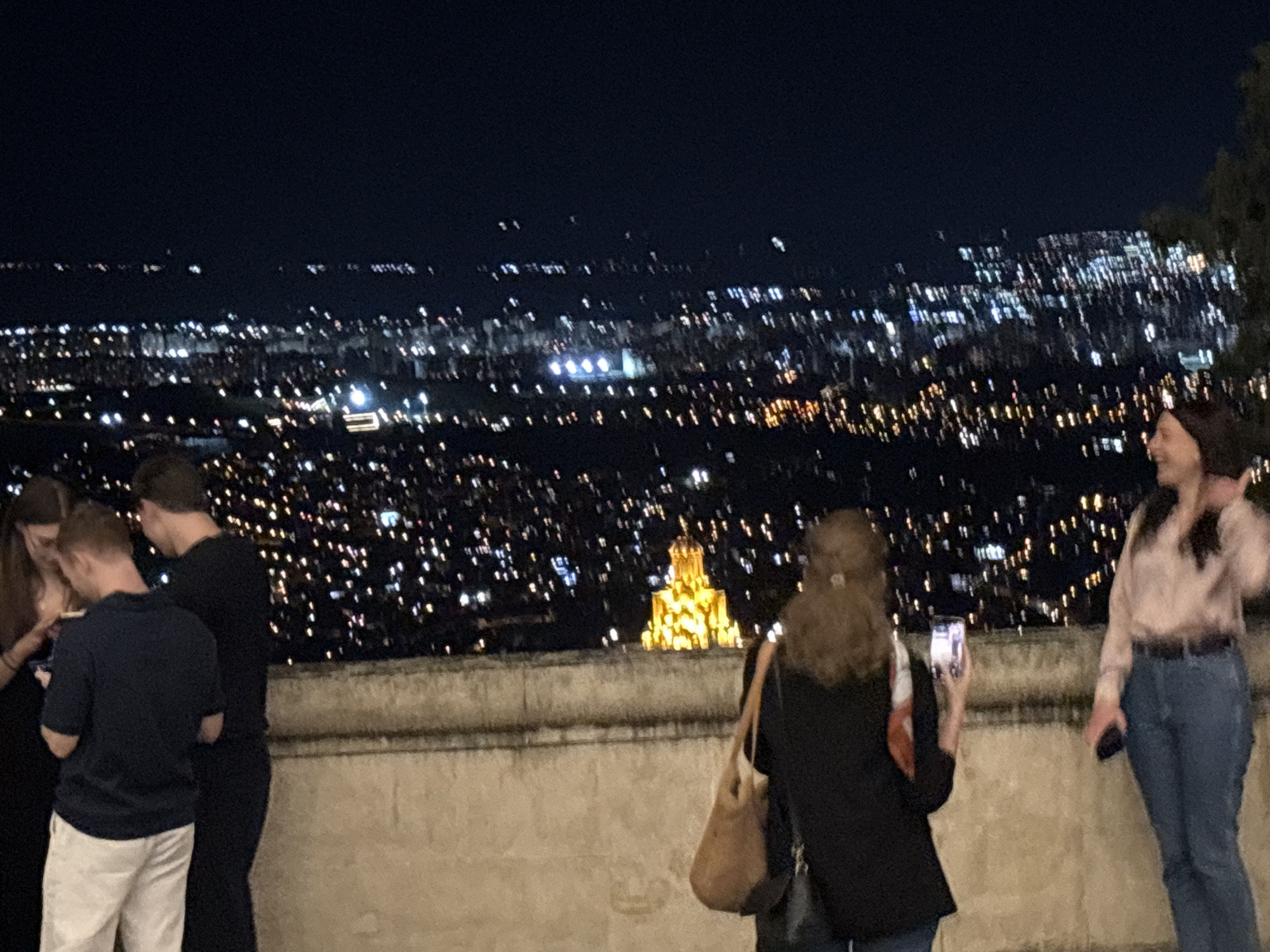The image shows a nighttime scene overlooking the city of Tbilisi, Georgia. The foreground features a group of people standing on a vantage point. On the left, three individuals are gathered, with two men and one woman visible. One man wears a dark blue polo shirt and light-colored pants, while the other wears a black long-sleeved shirt. The woman on the far left is mostly obscured. In the center, a woman with curly brown hair is holding a phone, seemingly taking a picture. She is wearing a black jacket over a patterned top and carrying a brown bag. To the right, a woman in a light pink blouse and blue jeans is also holding a phone.
The background is a sprawling cityscape illuminated by countless lights, indicating it is nighttime. A prominent, brightly lit building, possibly a church or monument, stands out in the middle ground, glowing with yellow light. The dark sky above is clear, with only a hint of distant lights suggesting a horizon. The perspective suggests the vantage point is elevated, offering a panoramic view of the city. The atmosphere is one of casual observation and enjoyment of the urban night view.