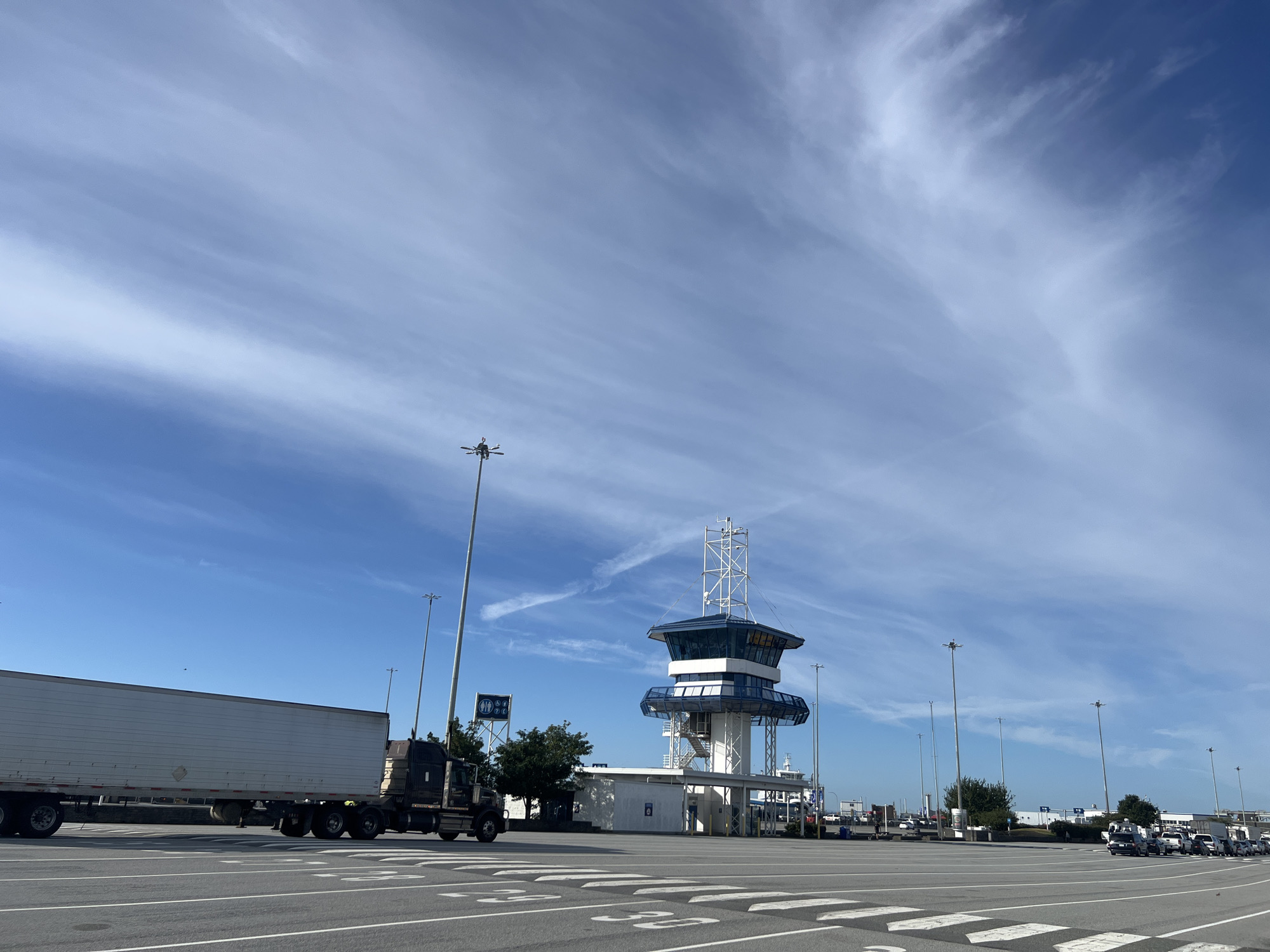 The image captures an outdoor scene at what appears to be a ferry terminal in Delta, Canada. The sky is a vibrant blue with wispy white clouds streaking across it, suggesting a clear and sunny day. Tall light poles are scattered throughout the paved area, which is marked with white lines indicating lanes and pedestrian crossings.
In the foreground and mid-ground, a large, white semi-trailer truck is parked, facing to the right. Behind the truck, a modern control tower with a blue-tinted glass observation deck stands prominently. The tower has a white, multi-story structure with a lattice-like antenna at its apex. To the left of the tower, a sign with blue circular icons is visible.
Further in the background, several other vehicles, including cars and trucks, are lined up, indicating that this is likely a waiting area or part of the ferry boarding process. The presence of a large ferry boat in the distance further supports the ferry terminal context. The overall atmosphere is one of a busy, operational transportation hub. No people are clearly visible, and the scene conveys a sense of organized activity. The time of day appears to be daytime, likely mid-morning or afternoon, given the bright sunlight and the shadows cast.