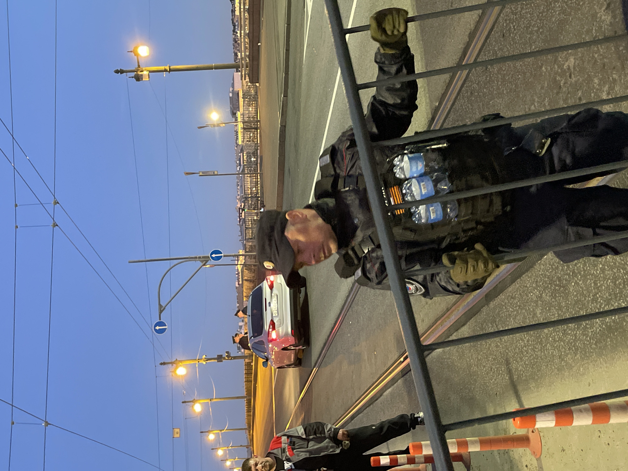 A man in a black uniform with a cap and gloves holds a portable metal barrier. He is wearing a vest with several bottles of water attached. Another man in a grey and red jacket stands to his left. In the background, a white car is stopped on a street lined with lampposts and overhead wires. The sky is a deep blue, indicating dusk or dawn. Orange and white traffic cones are visible on the left side of the image.