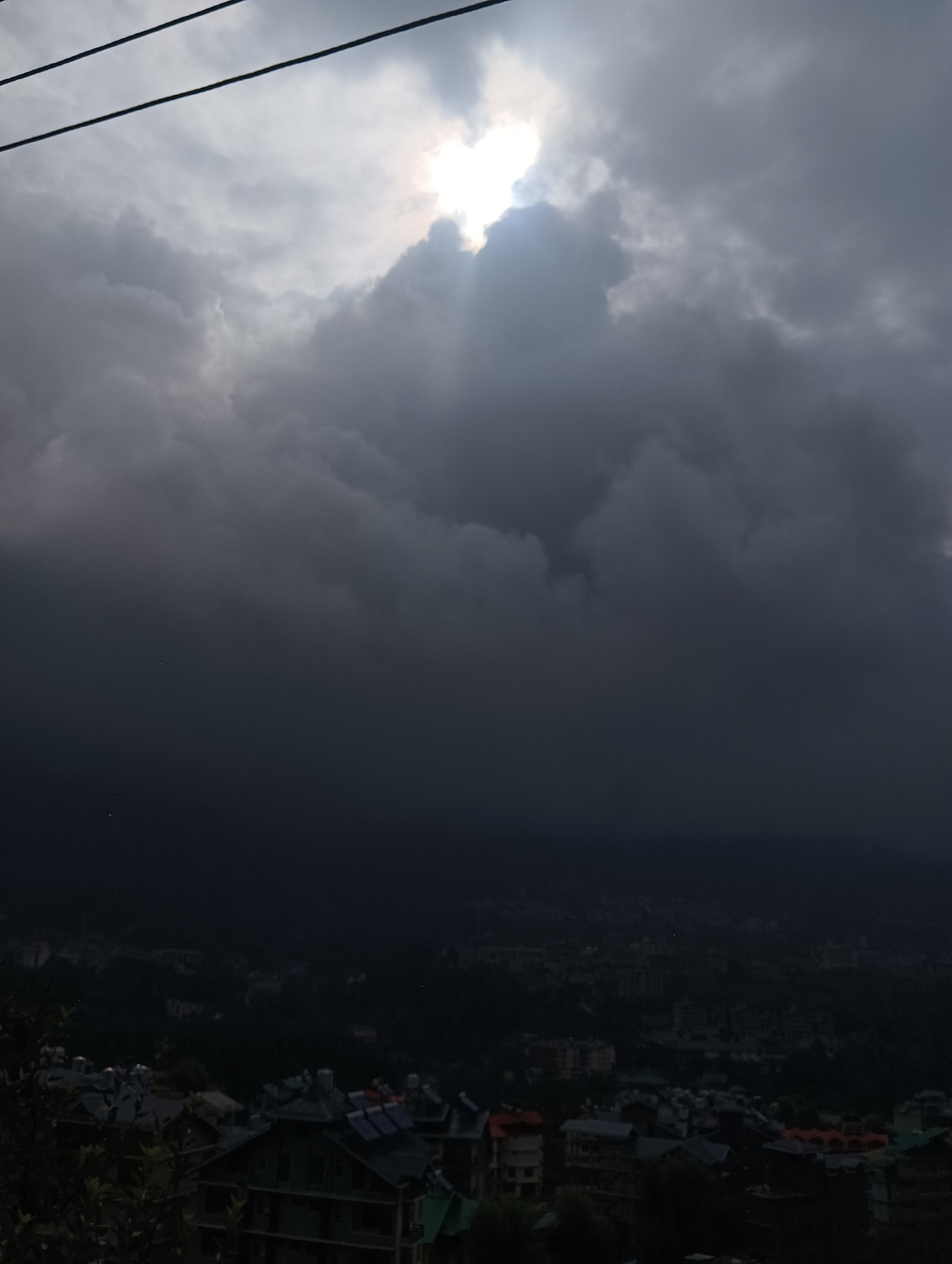 This is a photograph taken from a high vantage point overlooking the town of Siumsa, India, likely during a cloudy or stormy period. In the foreground, there are dark green foliage and parts of buildings with dark, sloped roofs, some of which appear to be equipped with solar panels. A few scattered structures with red roofs are also visible. The middle ground shows a densely populated urban area with numerous buildings, all under a very dark, heavy cloud cover that dominates the upper portion of the image. Sunlight is breaking through the clouds in a concentrated area towards the top center, creating a bright, diffused glow and casting visible light rays downwards. Three dark, thin power lines run horizontally across the top of the frame, just below the brightest part of the sky. The overall lighting is dim, with a sense of an impending storm or post-storm conditions.