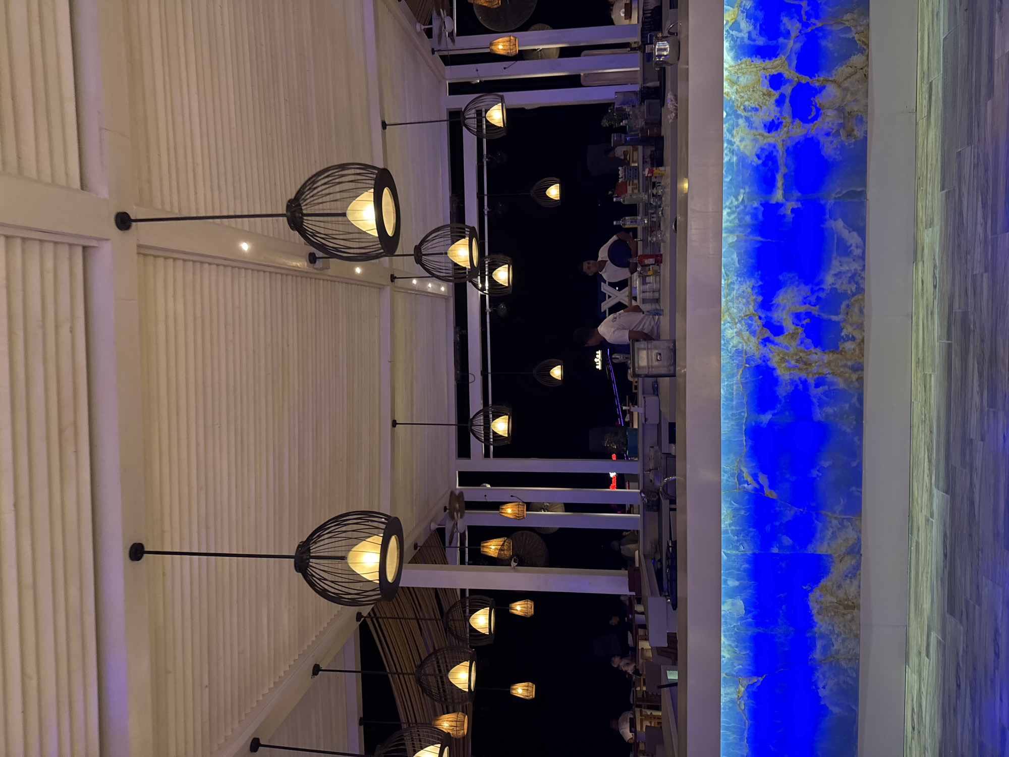 This is a photograph of an outdoor bar area at night in Unknown, Egypt. The bar is illuminated by several decorative pendant lights hanging from the ceiling, which has a slatted, white structure. In the foreground, the bar counter is visible, featuring a translucent blue onyx facade that is backlit, creating a vibrant glow. Behind the counter, several individuals, presumably staff, are working. One person in a white shirt appears to be serving drinks, and another in a white uniform is visible in the background. Bottles of alcohol and glassware are arranged on the counter. Beyond the bar, diners are seated at tables in the dim light. The overall ambiance suggests a lively, nighttime establishment.