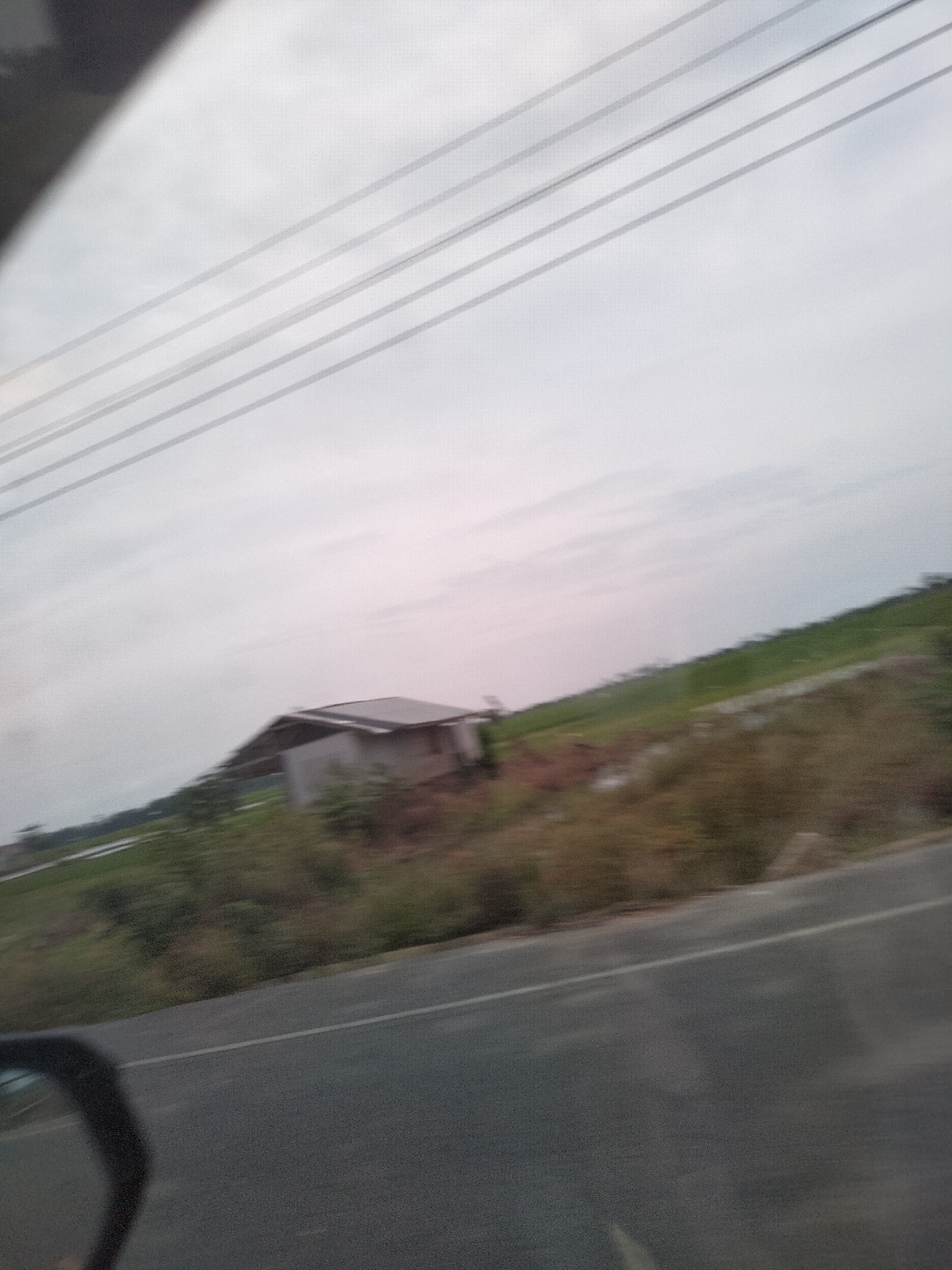 A blurry, wide-angle shot captures a rural scene in Matangpudeng, Indonesia. A small, light-colored building with a dark roof stands on a patch of elevated land covered in vegetation. The building appears to be a simple house or shed. In the background, a green field stretches towards the horizon under a cloudy sky. Power lines are visible diagonally across the upper portion of the image. In the foreground, a dark asphalt road with a white line marking the lane curves into the lower right corner of the frame. A portion of a dark car mirror is visible in the bottom left corner. The overall impression is a passing glimpse of a roadside view in a rural area.