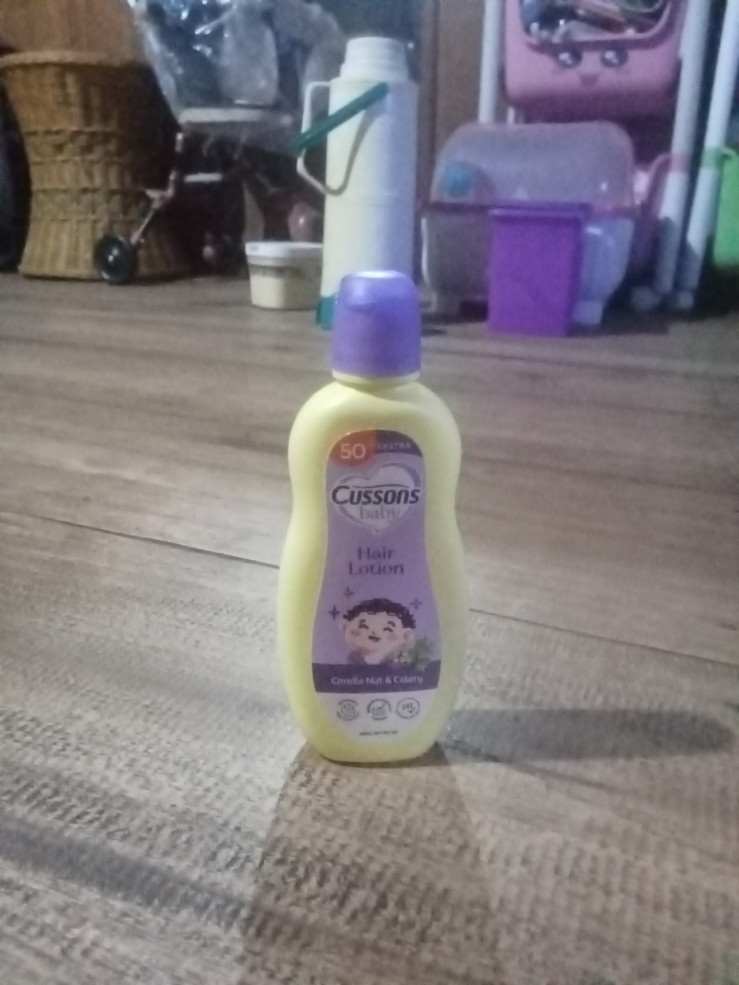 A bottle of Cussons Baby Hair Lotion with a purple cap is centered in the foreground, resting on a wooden floor. The bottle is light yellow and features a label with text and an illustration of a baby's head with curly hair. The background is blurred and shows various objects, including a woven basket, plastic containers, and a pink toy. The lighting suggests an indoor setting, possibly a residential area in Setia Jaya, Indonesia.