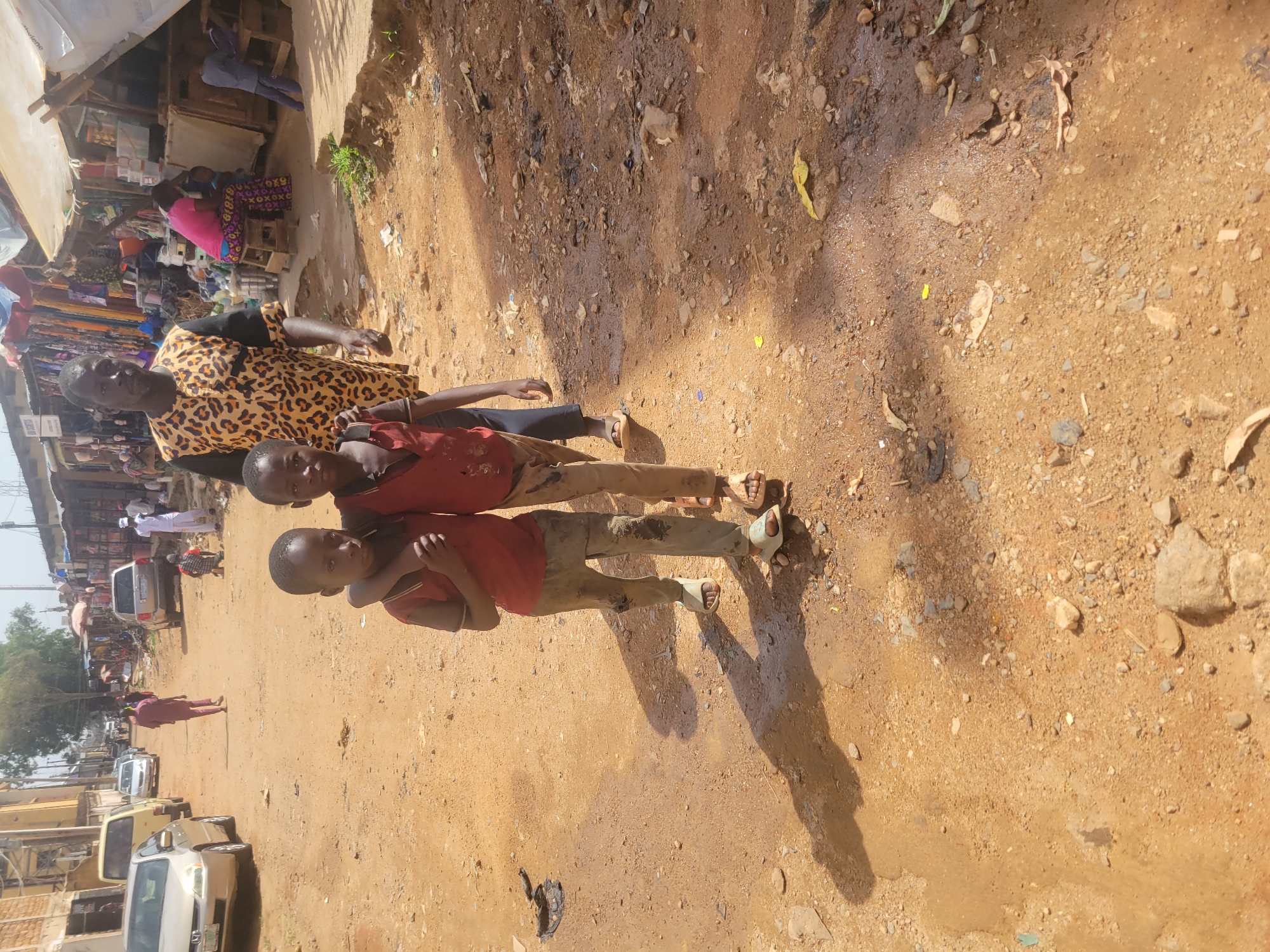 Three boys stand embraced on a dirt road in Jos, Nigeria, with their shadows cast beside them. The boys wear casual clothing, including red shirts and sandals. A man in a leopard-print shirt stands to their side. A bustling market is visible in the background, with shops, stalls, and pedestrians. Cars are parked on the opposite side of the road. The ground is dry and dusty, with scattered rocks and debris.