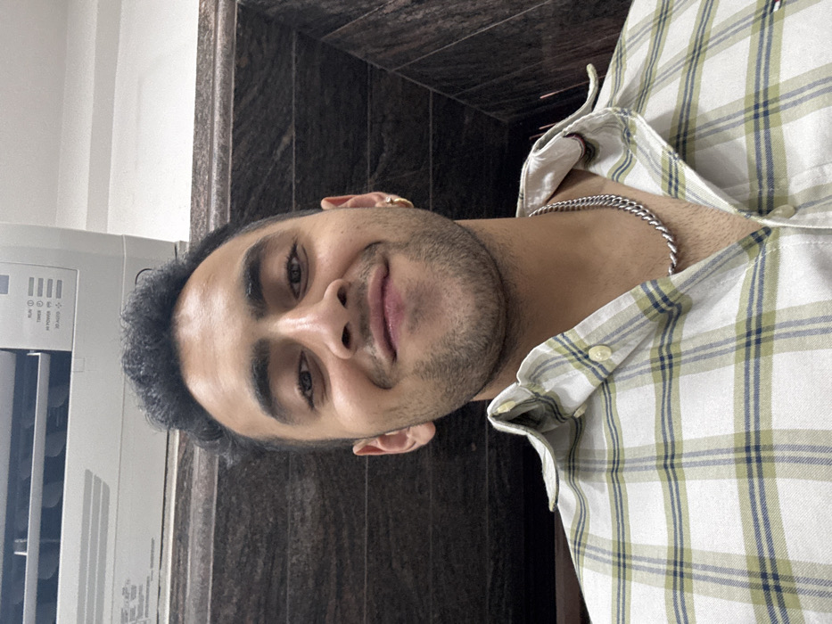 A young adult male is depicted in a close-up, chest-up indoor shot, positioned centrally and looking directly at the camera with a slight, closed-mouth smile. He has short, dark, curly hair and stubble on his face. He is wearing a button-up plaid shirt featuring a pattern of white/off-white, light green, and dark blue/black intersecting lines. A silver-toned curb chain necklace is visible around his neck, and a small gold hoop earring is present in his right ear (viewer's left).
The background consists of an interior setting. Directly above the subject's head is a white, wall-mounted split-system air conditioning unit with visible function indicators, including "RUN," "TIMER," "HI POWER," "SLEEP," and "3D AUTO." Below the air conditioning unit and behind the subject, a dark, polished surface, consistent with stone or marble paneling with horizontal striations, covers the wall. The upper right portion of the image reveals a white ceiling and an angled dark wall or structural element. The specific location within the city of Kolkata, India, cannot be determined from the visual information provided beyond it being an interior space, likely a residence or commercial establishment.