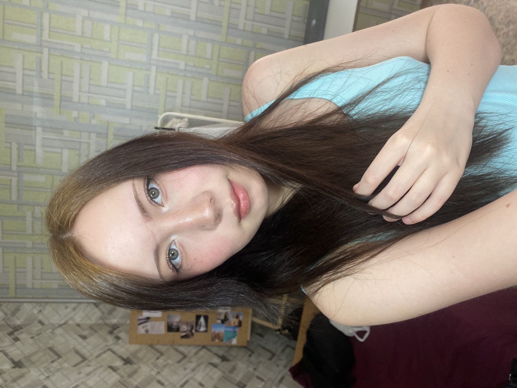 A close-up, rotated, selfie-style image features a young woman with light skin and long, dark brown hair with blonde streaks. She has light eyes and is wearing a light blue tank top. The background is a room interior. The wall behind her is decorated with geometric wallpaper in shades of green, grey, and white. There's a collage of photos visible on a corkboard in the lower left corner of the image. A bed with dark purple sheets is partially visible in the lower right corner. The location is Unknown, Unknown.