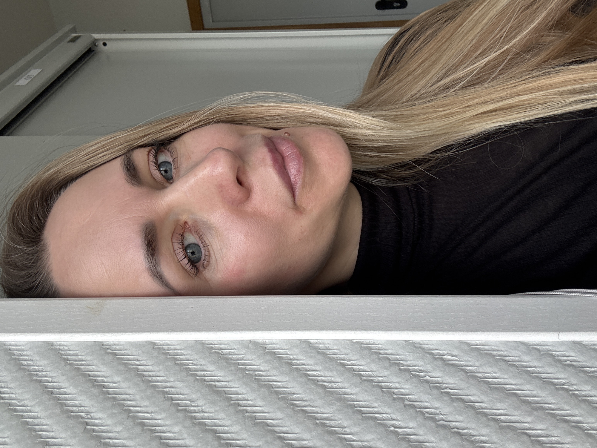 A blonde woman is pictured lying on her side, looking directly at the viewer with blue eyes and a calm, neutral expression. Her head is positioned towards the left side of the frame, and her long, straight blonde hair flows behind her. She has fair skin, visible eyelashes enhanced by mascara, and light pink lips. A subtle, clear stud or small blemish is visible on her upper lip area. She is wearing a dark, possibly black, ribbed or textured top.
The setting appears to be indoors. In the immediate foreground, a white or light gray textured surface, likely a mattress or bed cover, displays a distinct diagonal corrugated pattern. Above this patterned surface is a smooth, light-colored frame or edge, presumably part of a bed or headboard. The background consists of a plain, light gray surface, which could be a wall, the underside of a shelf, or a piece of furniture. A small, partially obscured label with faint text, possibly "100," is visible on this background surface in the upper left corner. The image is rotated, giving the impression of the woman lying on a vertical surface. The lighting is soft and even, suggesting indoor ambient light. The location is contextualized as Chaikovsky, Russia, though no specific visual cues within the image support this.