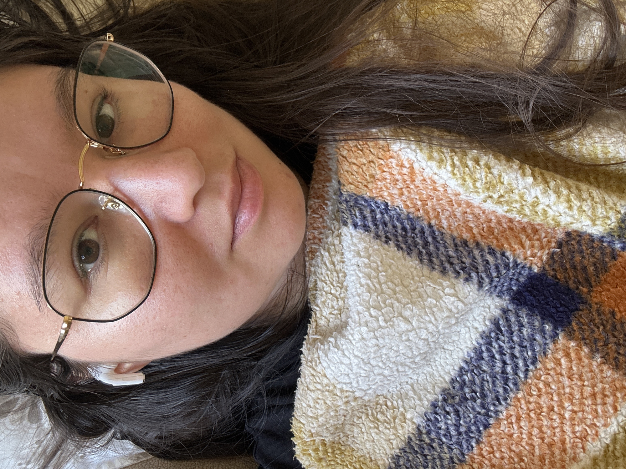 A woman lies down with her head resting on a plaid blanket with a white, orange and blue pattern. She is wearing glasses with a thin black frame and gold accents, and has long dark brown hair. There is a white object (possibly an earbud) visible near her left ear. The location is not discernible from the image.
