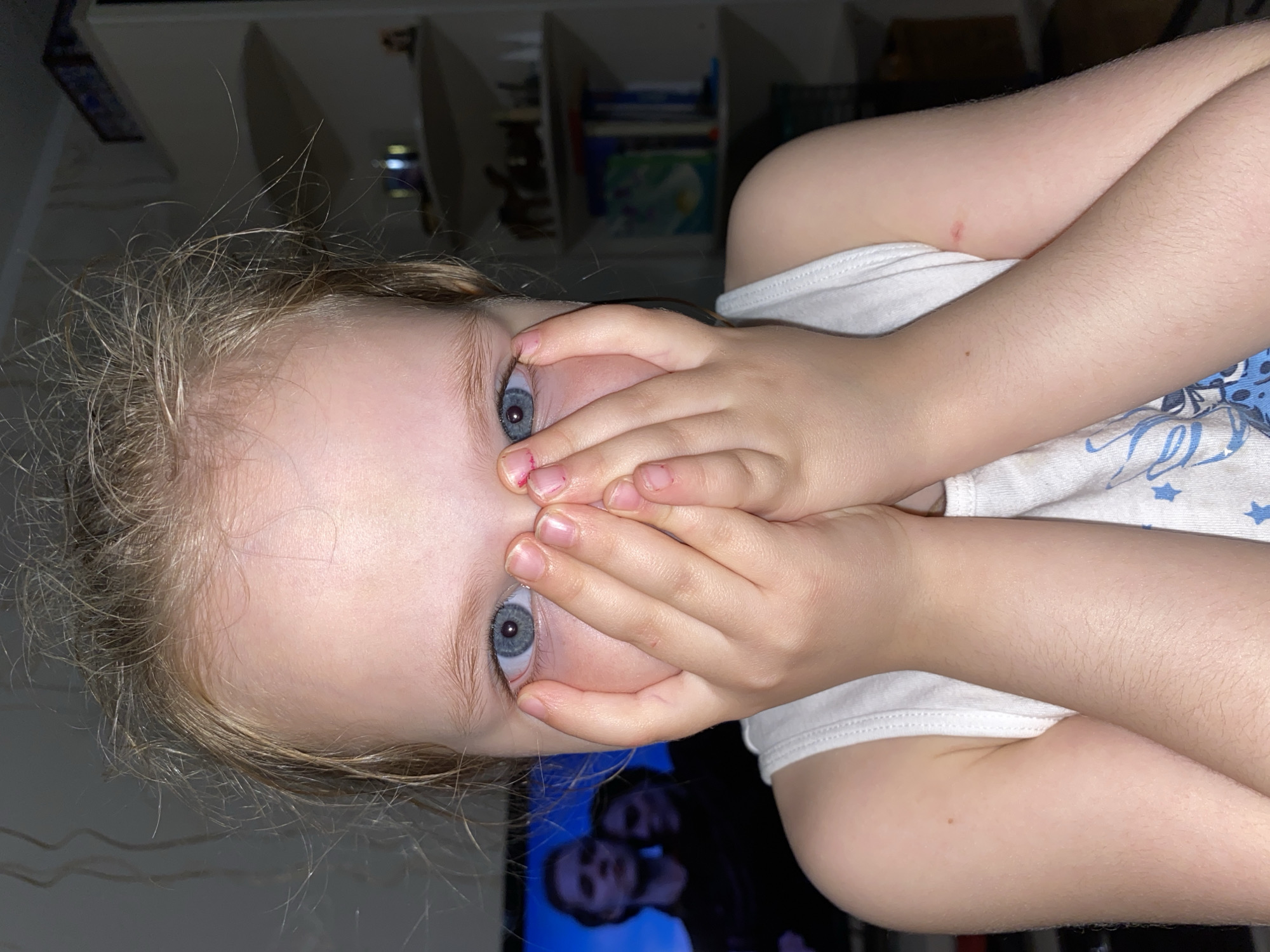 A young, light-skinned child with blond hair and blue eyes is covering their mouth with their hands. The child is wearing a white tank top. The background includes a wall unit with shelves and books. A television screen is also visible, showing two figures on it. No information suggests that the image was taken in Votkinsk, Russia.