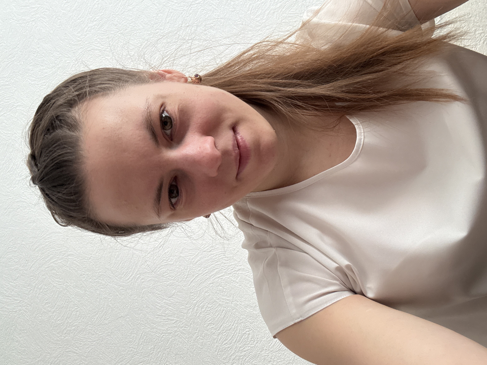 A young woman is pictured in a close-up head and shoulders shot, slightly angled. She has light brown hair, partially braided back from her forehead, and hazel eyes. She wears a light-colored, short-sleeved blouse. The background is a plain, textured white wall. The location is not discernible from the provided image. The absence of specific identifying details precludes any definitive determination of the image location in Chaikovsky, Russia, or any other location.