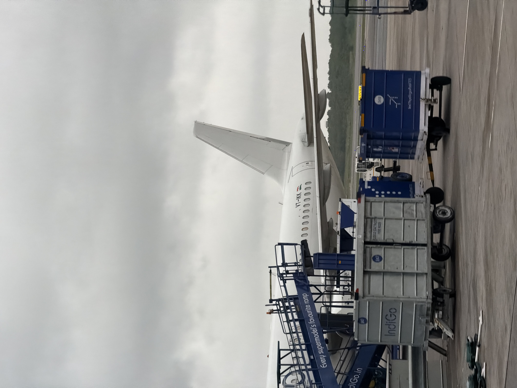 An overcast sky hangs above a white IndiGo airplane parked on the tarmac at Rajiv Gandhi International Airport in Hyderabad, India. A blue boarding ramp with the text "Every supermodel's favourite ramp" is positioned adjacent to the aircraft's fuselage. Several luggage carts, one grey with "IndiGo" branding and another blue with the IndiGo logo, are visible in the foreground. The airport tarmac is paved, and a line of trees can be seen in the distance. The scene is static and devoid of any visible individuals.