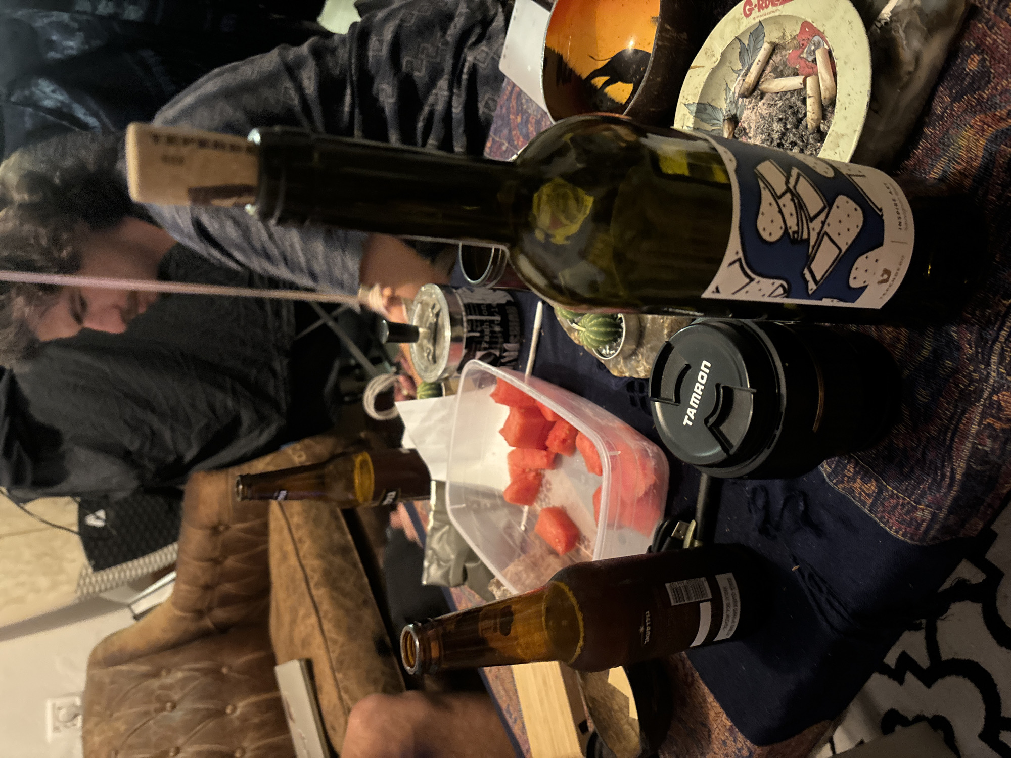 The scene is indoors, cluttered with various objects on a table covered with a patterned cloth. A person with curly hair is partially visible, wearing a dark shirt. A wine bottle with a cork is prominent, featuring a label with blue and white geometric designs. A Tamron lens cap sits nearby. A plastic container holds cut pieces of watermelon. An ashtray filled with cigarette butts is also visible. Two beer bottles are on the table as well. The location cannot be definitively confirmed as Tel-Aviv, Israel based on the visual information provided.