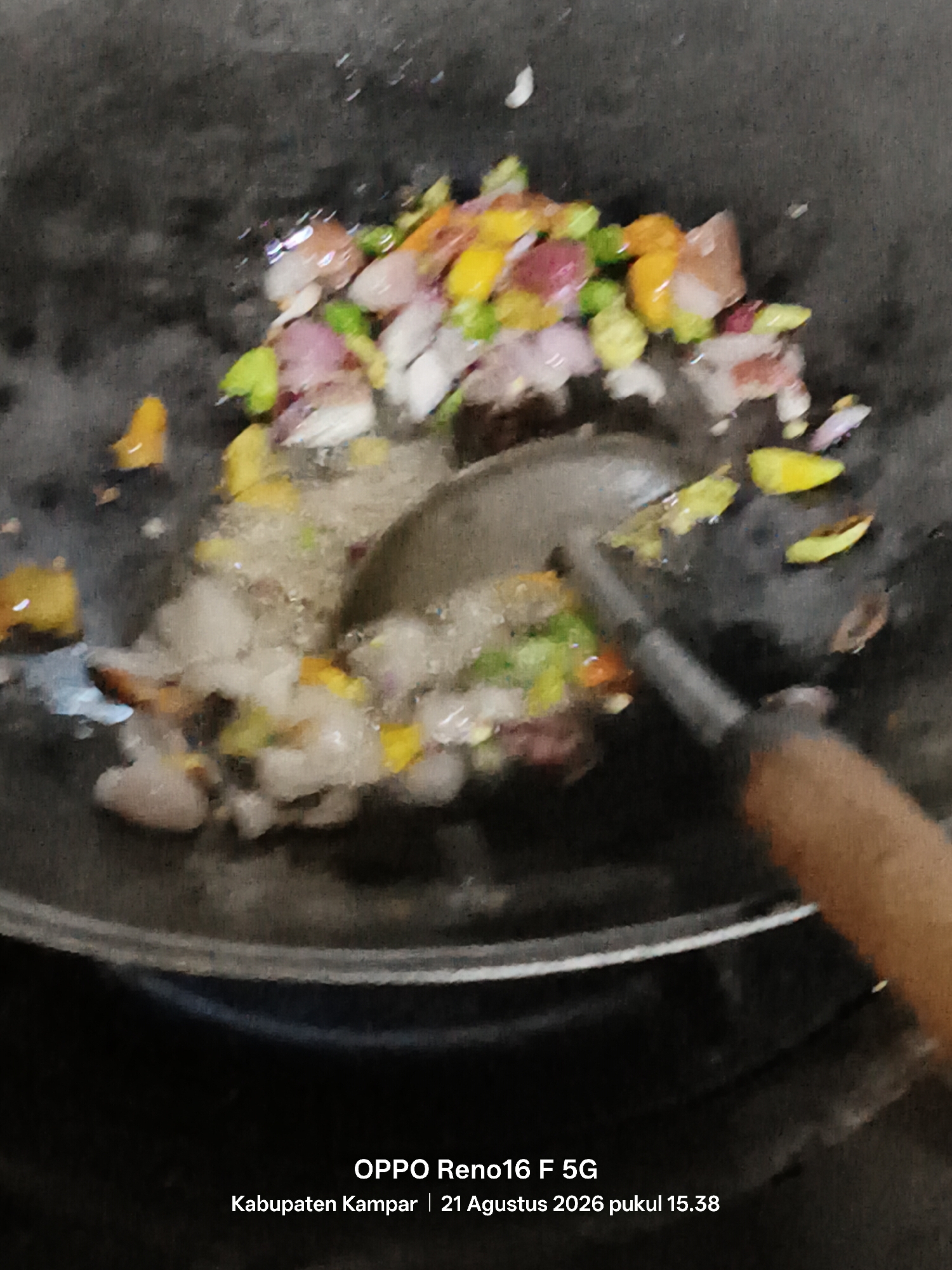 The image shows a close-up, slightly blurry overhead view of food being stir-fried in a dark, non-stick wok. Chopped vegetables, including what appear to be red onions, green peppers, and yellow bell peppers, are mixed with some translucent ingredients, possibly garlic or ginger, and a small amount of liquid, suggesting they are being sautéed. A metal cooking utensil with a wooden handle is partially visible, actively stirring the ingredients. The background is dark and textured, indicative of the wok's surface. The bottom of the image contains a watermark indicating "OPPO Reno16 F 5G Kabupaten Kampar | 21 Agustus 2026 pukul 15.38," suggesting the photo was taken in Kampar Regency, Indonesia, on August 21, 2026, at 15:38. There is no indication of the city of Penghidupan.