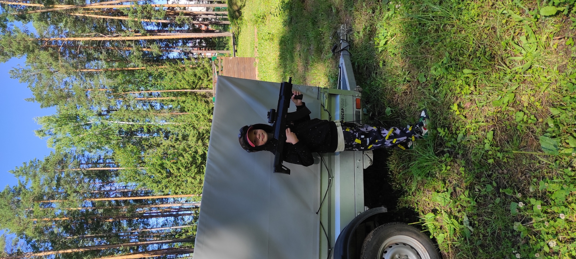 A young child, wearing a black hoodie with white speckles, a red baseball cap, and patterned trousers, stands in front of a gray trailer. The child is holding a black toy rifle with a scope. The child is also wearing a headset with lights, suggesting they are participating in a game like laser tag. The trailer appears to be parked on a grassy area with scattered vegetation. In the background, a dense forest of tall pine and birch trees is visible under a clear blue sky. A wooden structure, possibly part of a playground or activity area, is present to the right of the trailer. The overall setting is outdoors, likely in a park or recreational area in Noviy, Russia, given the presence of typical Russian forest vegetation.