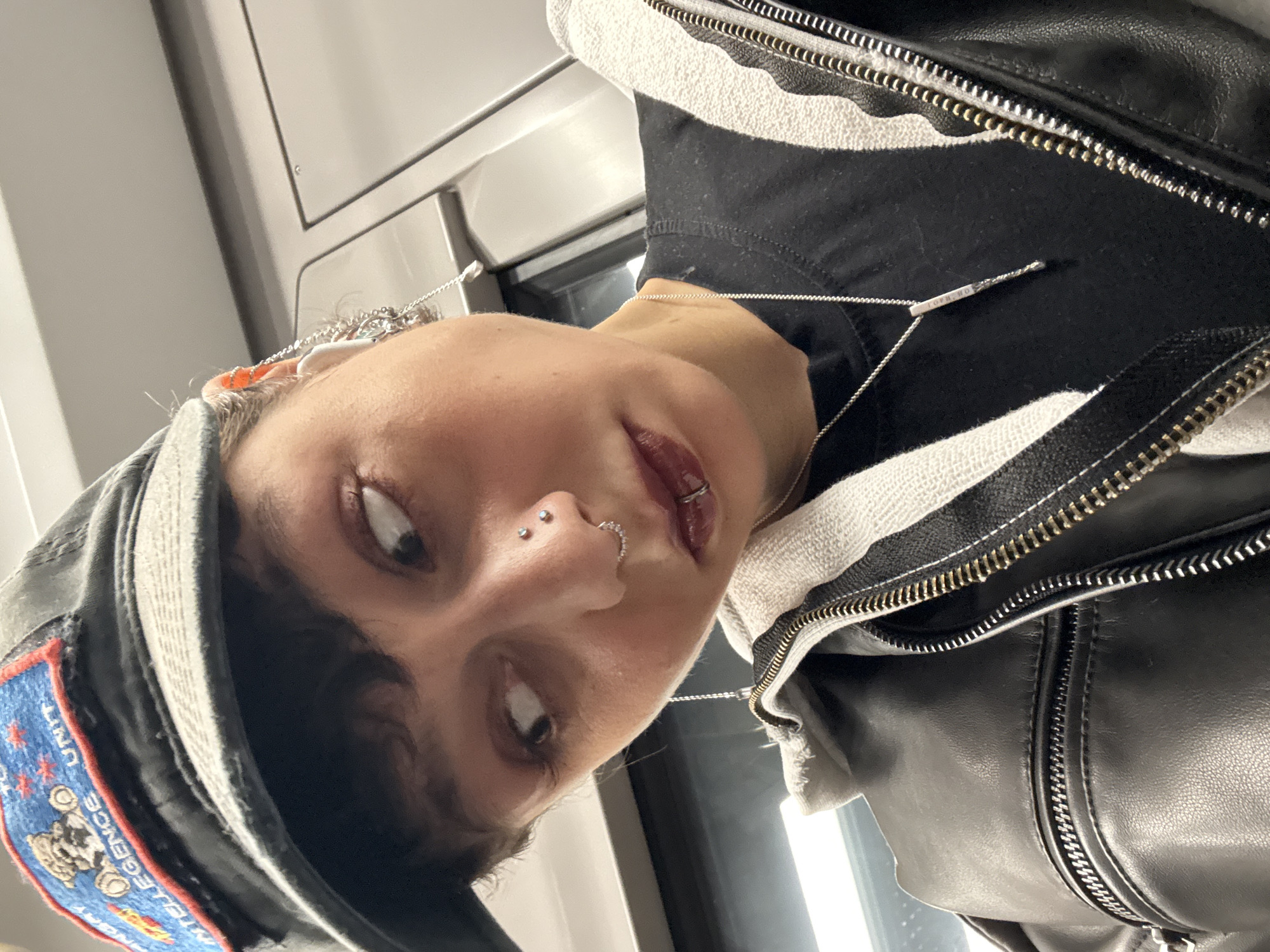 A close-up, slightly angled selfie of a person with dark hair, light skin, and several facial piercings (nostril, septum, and labret). The person wears a dark baseball cap with a patch (likely military or unit-related) on the side, a black t-shirt, and a black leather jacket with visible zippers. A thin silver necklace with a small pendant is also visible. The background appears to be the interior of a train or subway car, suggested by the neutral-colored walls and window frame visible in the background. There are no visible signs indicating this is Moscow, Russia.