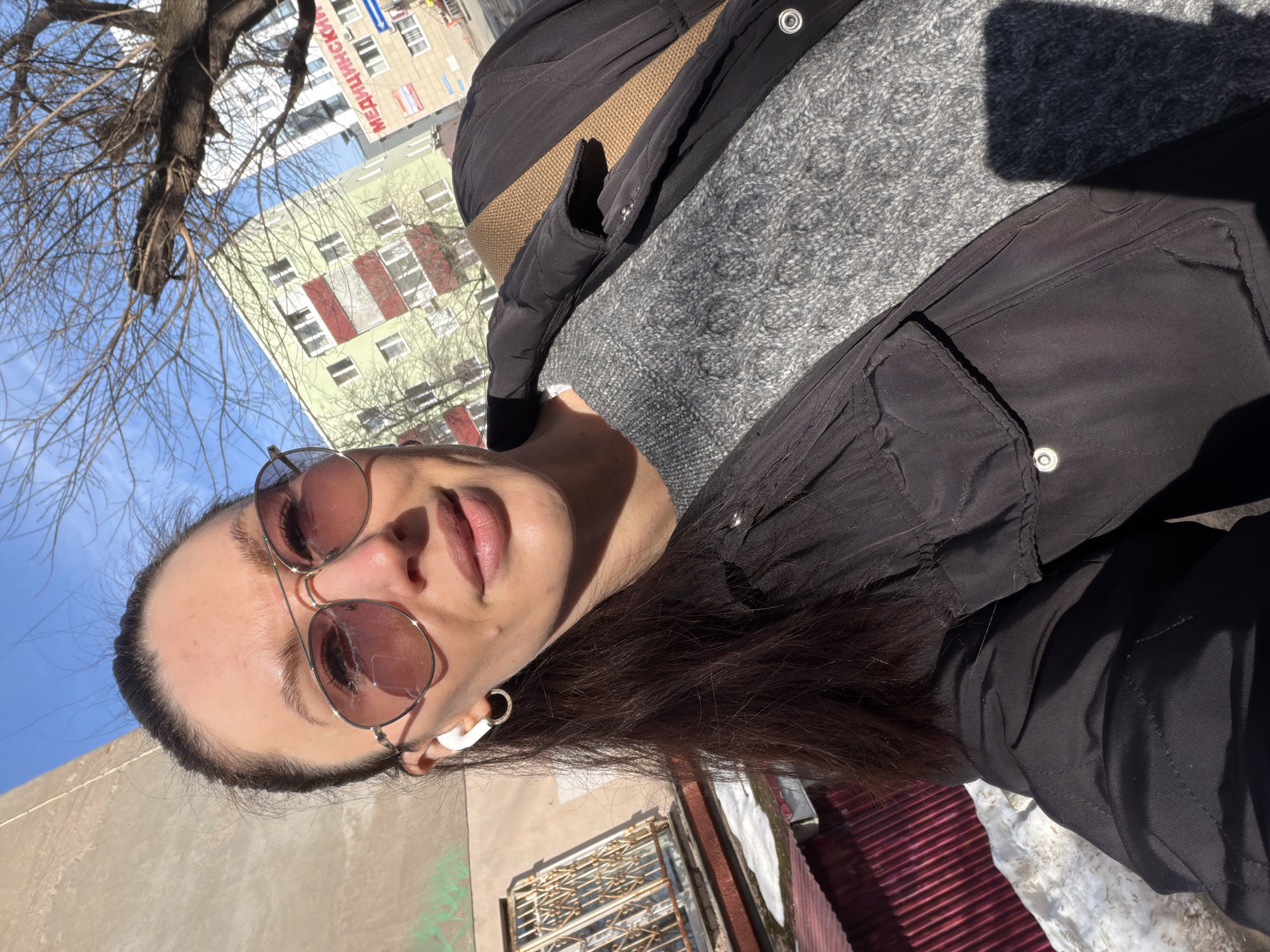 A woman wearing sunglasses and a black jacket stands outside, taking a selfie. She has brown hair and is wearing a gray sweater underneath the jacket. She also has a white earbud in her ear. Behind her, a building with the word "МЕДИЦИНСКИЙ" (Medical) is visible, suggesting the location is Perm, Russia. A bare tree is in the upper left corner. A low wall and metal structure are at the bottom right. The sky is blue.
