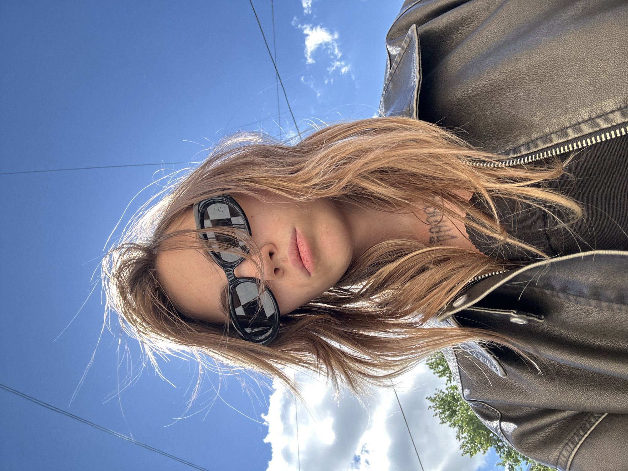 A young woman with blonde, wind-blown hair is seen from a low angle against a bright blue sky with scattered white clouds. She is wearing black, oval-shaped sunglasses with a checkered reflection and a black leather jacket. Her expression is neutral, and her lips are slightly parted. A tattoo is visible on her neck. The background includes visible power lines and a hint of green trees at the bottom left. The scene appears to be outdoors in an urban environment, possibly Moscow, Russia, given the presence of power lines and general urban landscape.
