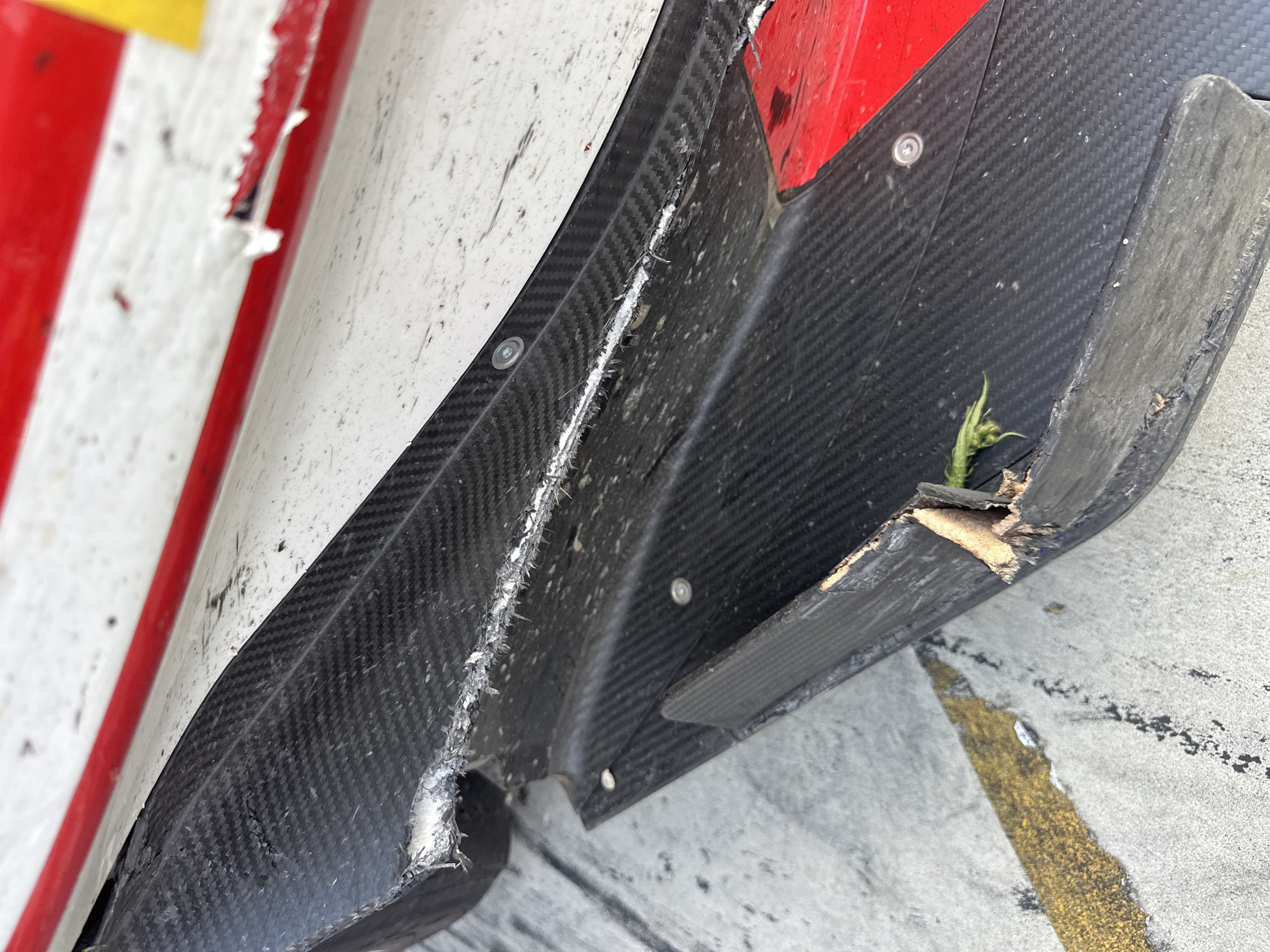 The image shows damaged carbon fiber bodywork from a race car, with a small weed growing out of a broken section. The bodywork is red and white, and is attached with silver rivets. The background is a concrete surface with a yellow line. The photograph was likely taken at the Autodromo Nazionale Monza (Monza Circuit) in Monza, Italy, as evidenced by the concrete surface and yellow line, which are common features of race tracks.